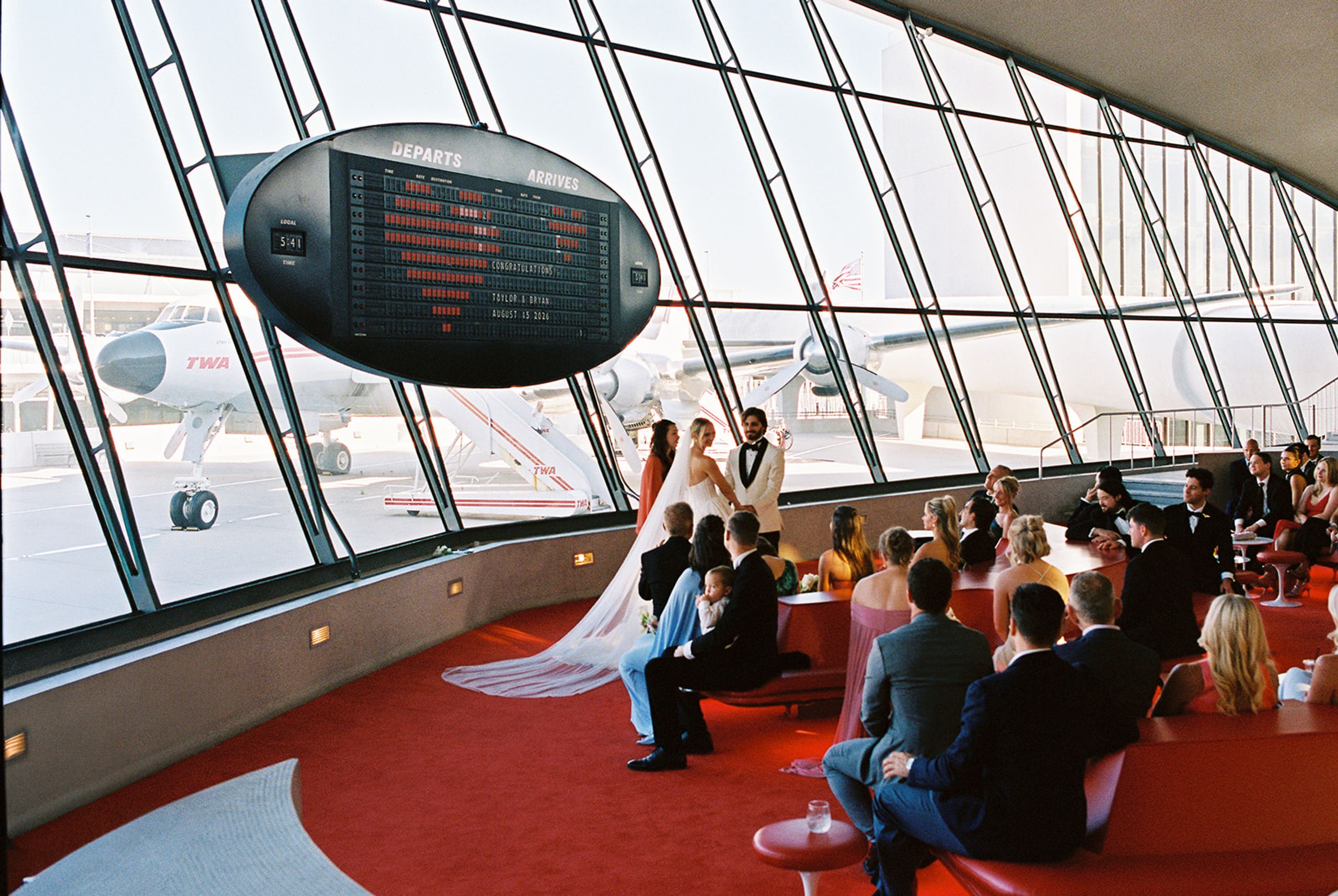 Wedding ceremony beneath the split-flap departures board at the TWA Flight Center with a vintage TWA plane outside