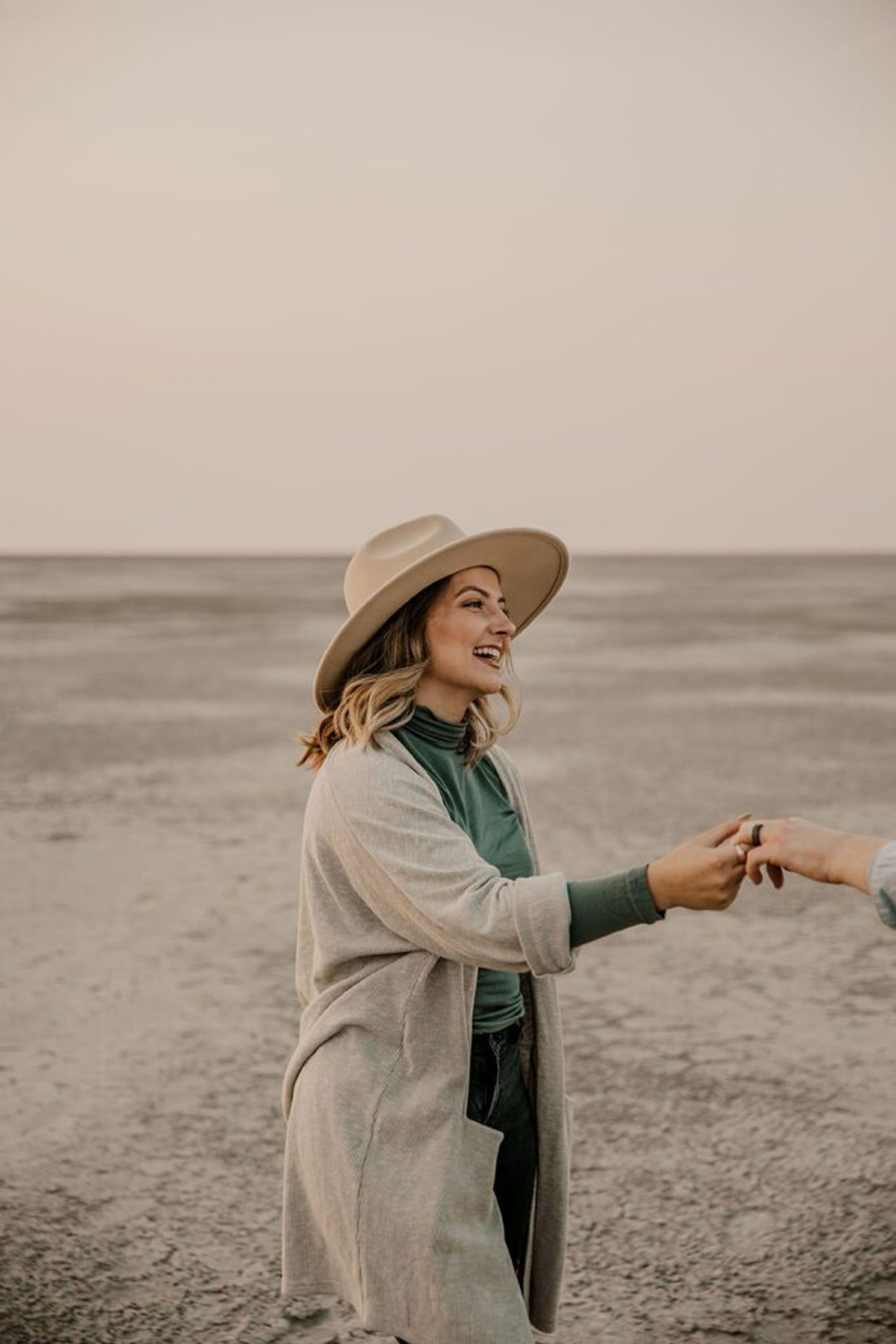 Woman smiling and holding hands with her partner during an engagement photoshoot in a desert setting, wearing a hat and casual fall attire.