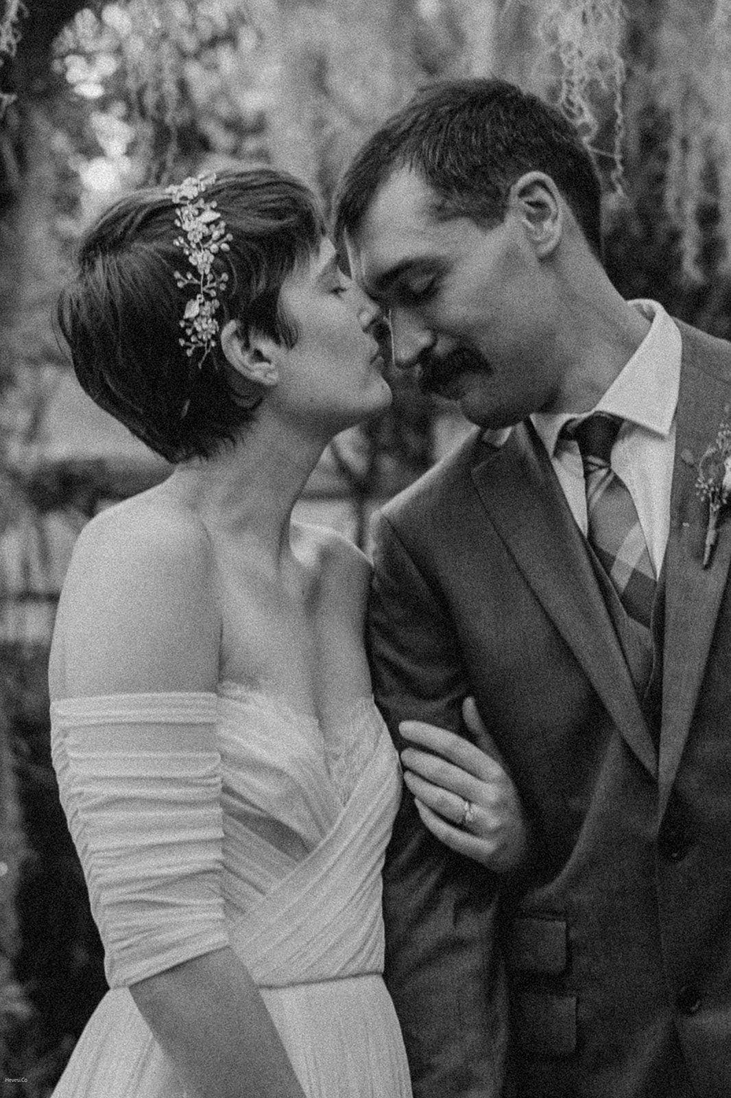A bride and groom share a tender moment, with the bride in an off-shoulder dress and the groom in a suit, standing closely together in a garden setting.