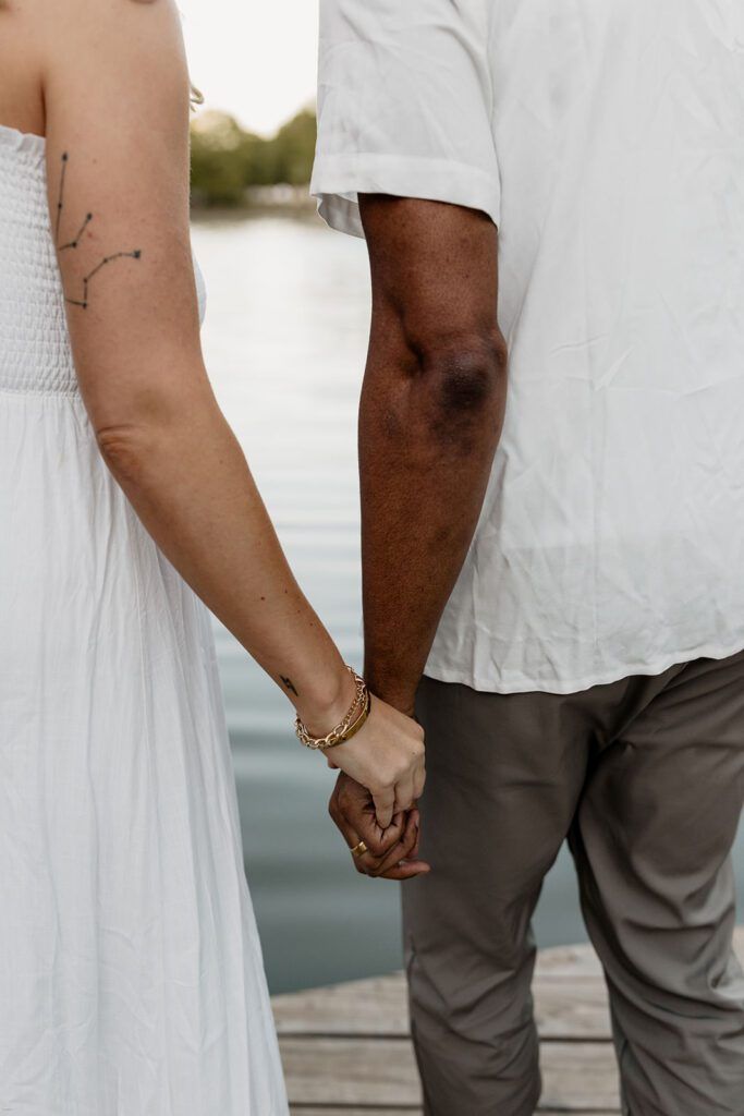 Engaged couple holding hands by the water, wearing casual summer outfits during their engagement photoshoot.