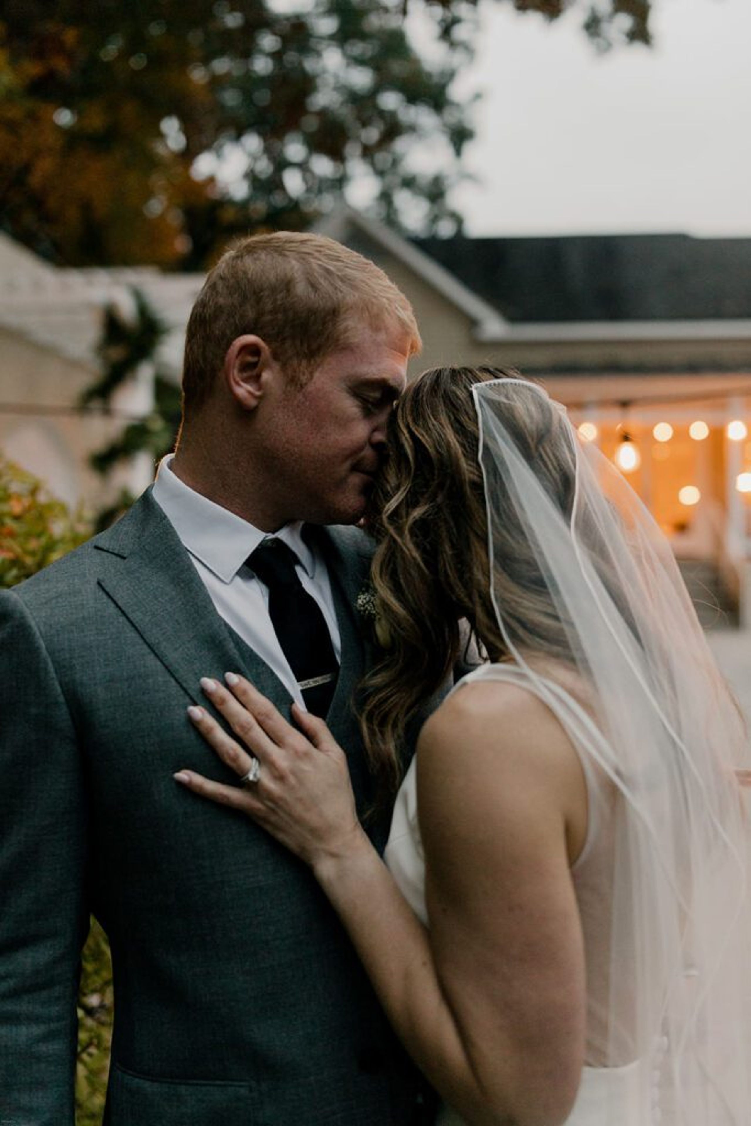 Scott gently kisses Deb's forehead as they embrace in a tender moment outside 173 Carlyle House, with warm evening lights glowing in the background.