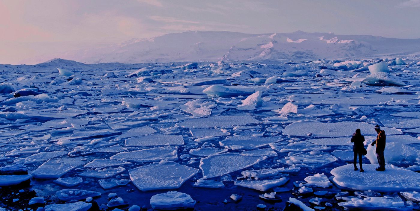 Two people stand on a small sheet of ice surrounded by a many other small sheets of ice on a body of water_