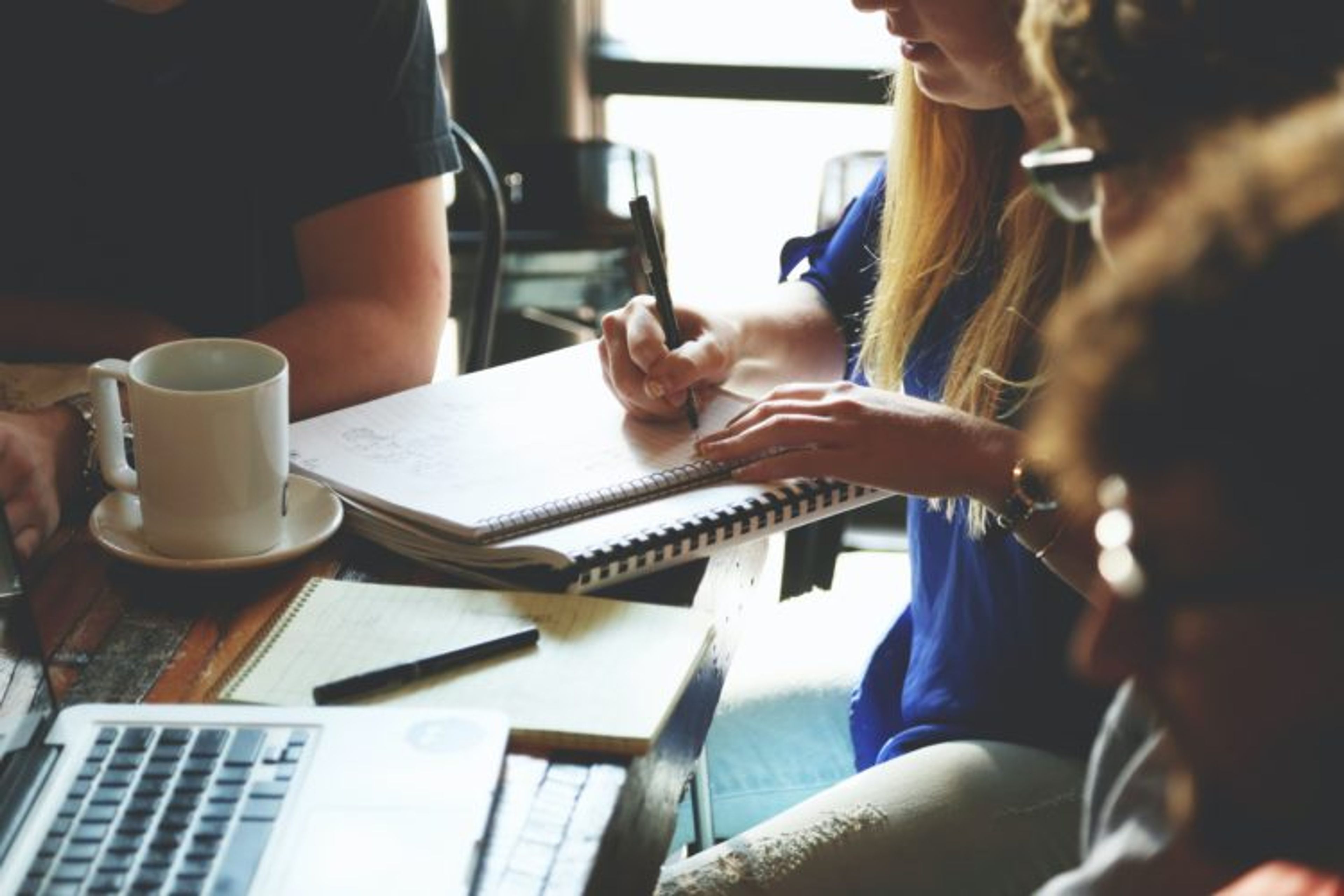 Four people seated around a table. One person writes in a notebook. An open laptop is in front of another person_