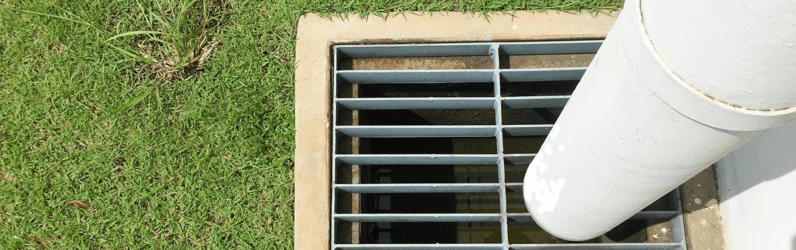 A white PVC downpipe discharging into a grated stormwater pit set in a lawn