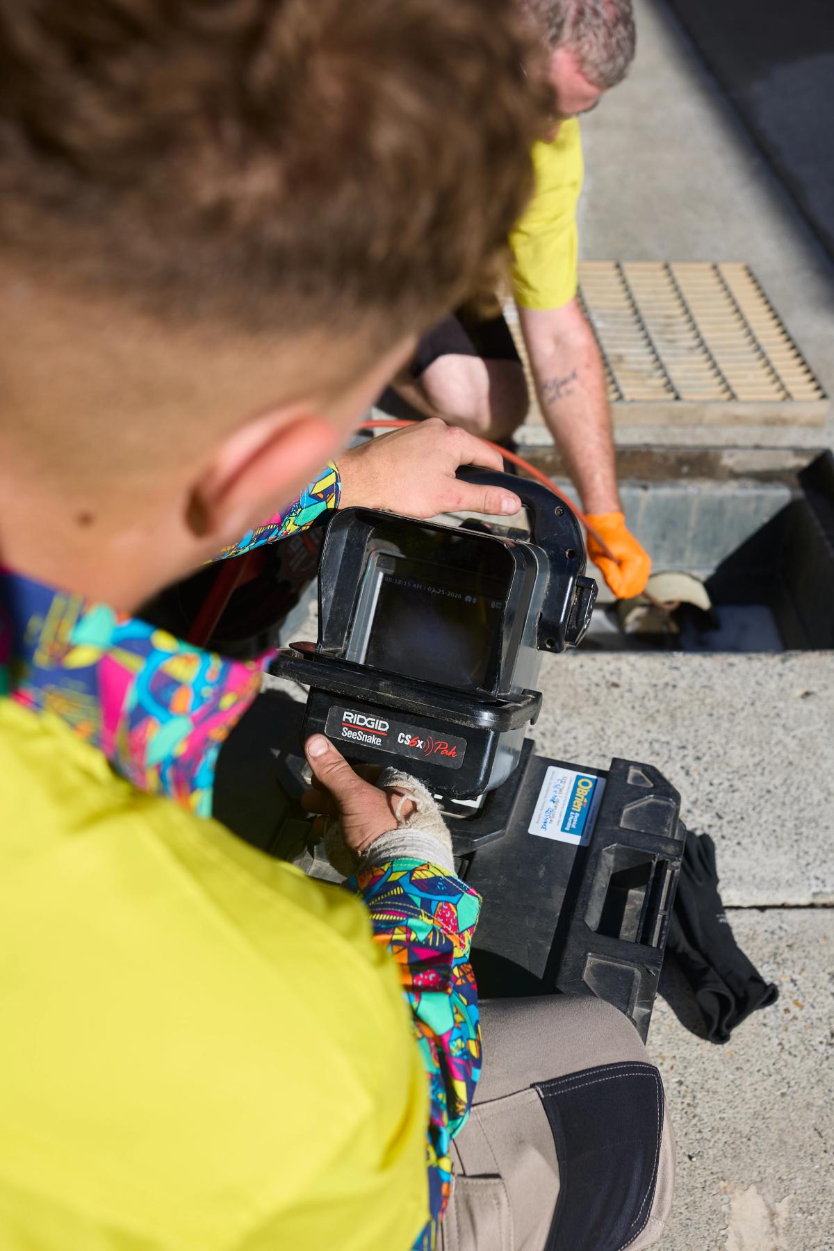 An O’Brien plumber holding a drain camera monitor while a colleague works at the drain