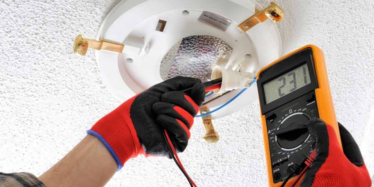 Gloved hands testing a ceiling light fitting with a multimeter