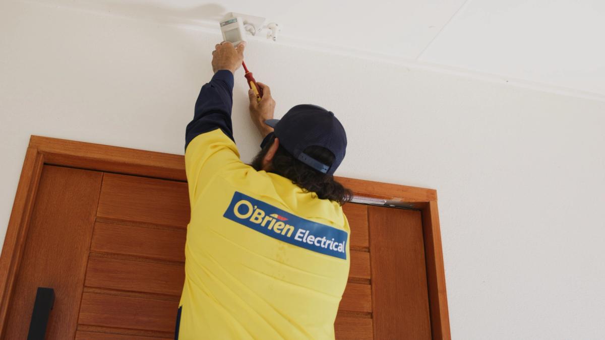 An O’Brien electrician fitting a ceiling-mounted sensor above a doorway