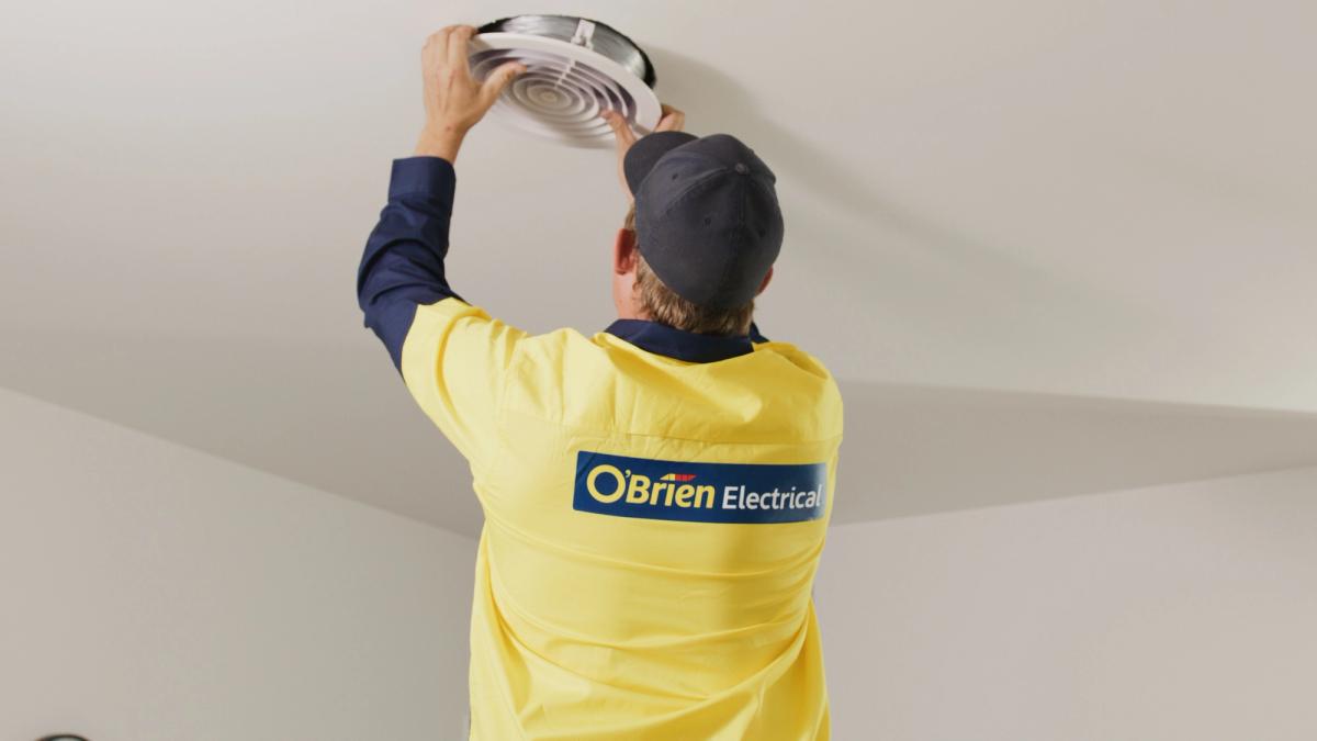 An O’Brien electrician reaching up to a round ceiling air conditioning vent
