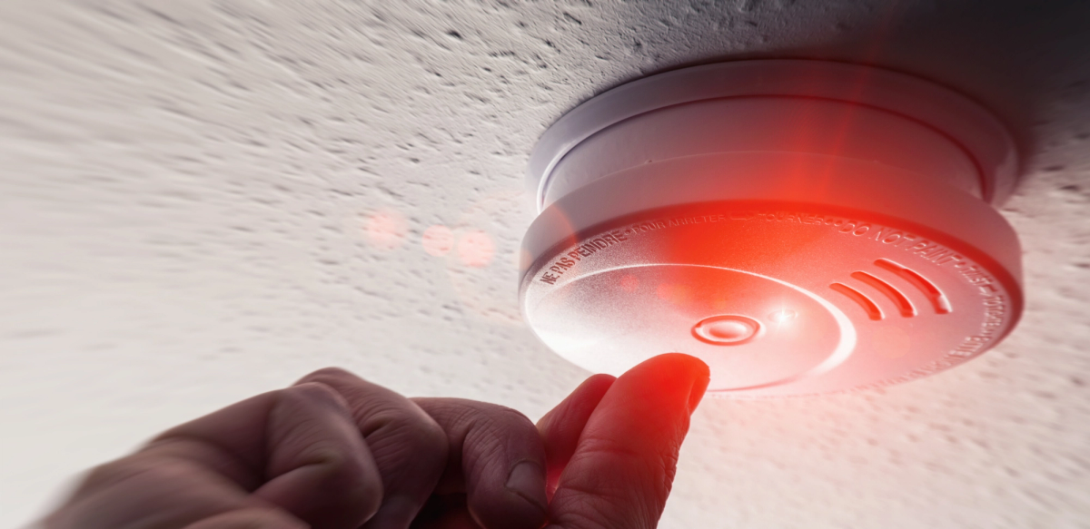 A finger pressing the test button on a ceiling-mounted smoke alarm