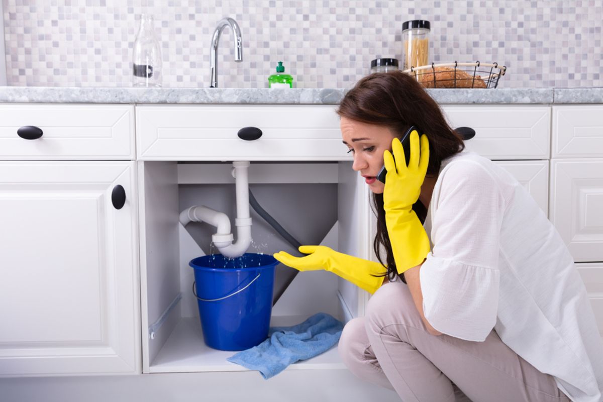 A person on the phone beside a leaking sink, catching water in a bucket