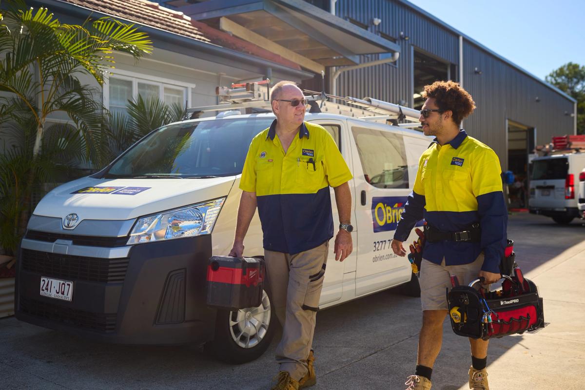 Two O’Brien technicians carrying tool bags past a branch van