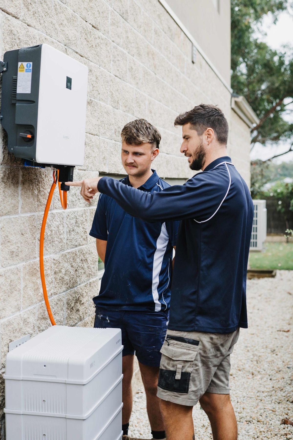Two technicians beside a wall-mounted inverter during a home battery installation