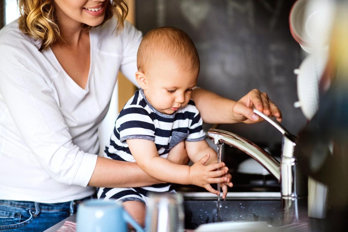 A parent holding a baby beside a running kitchen tap