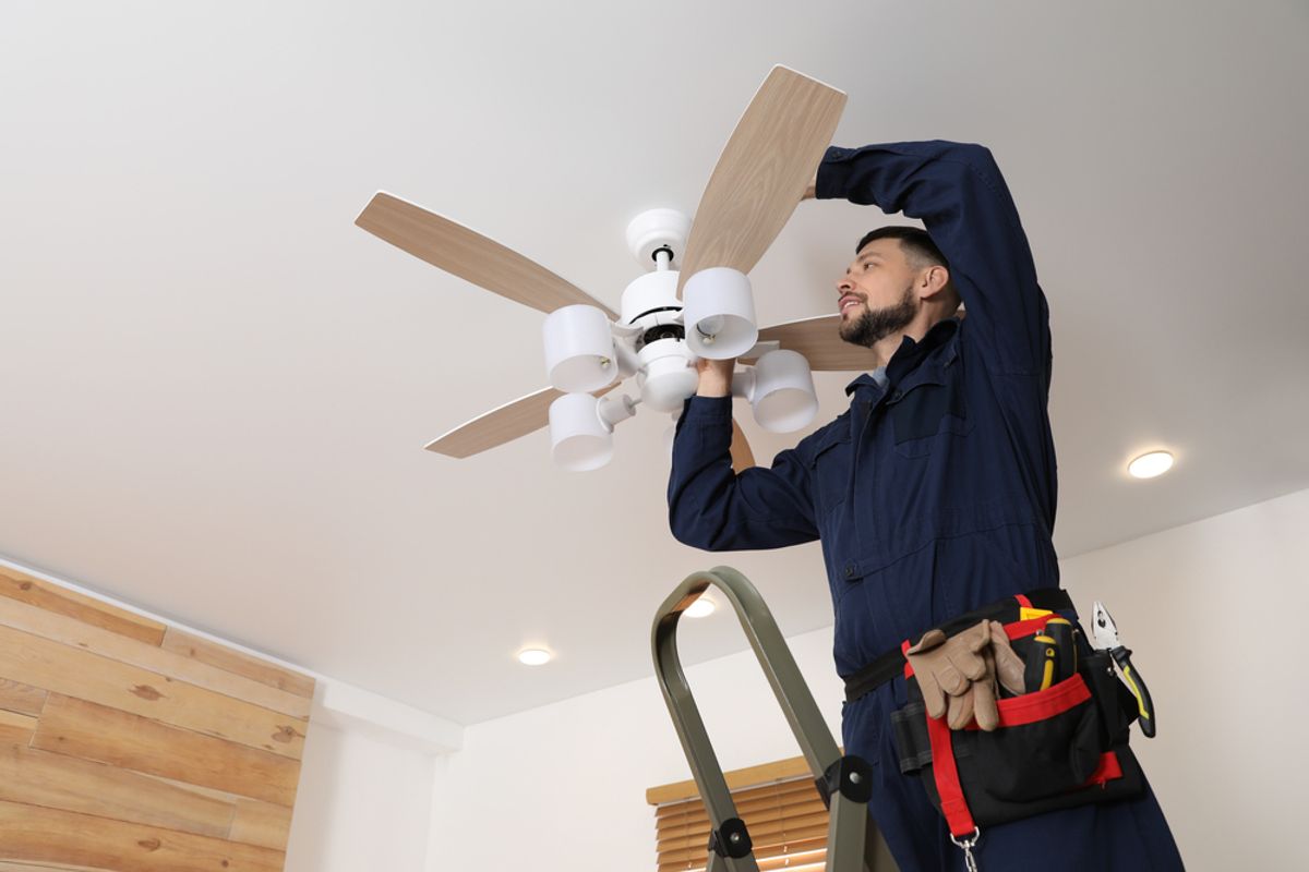 An electrician on a ladder fitting a ceiling fan with a light