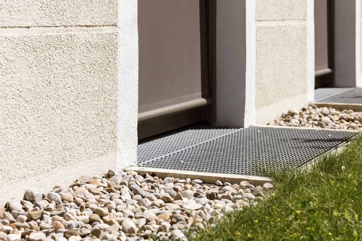 A grated stormwater channel drain set in gravel beside a rendered wall
