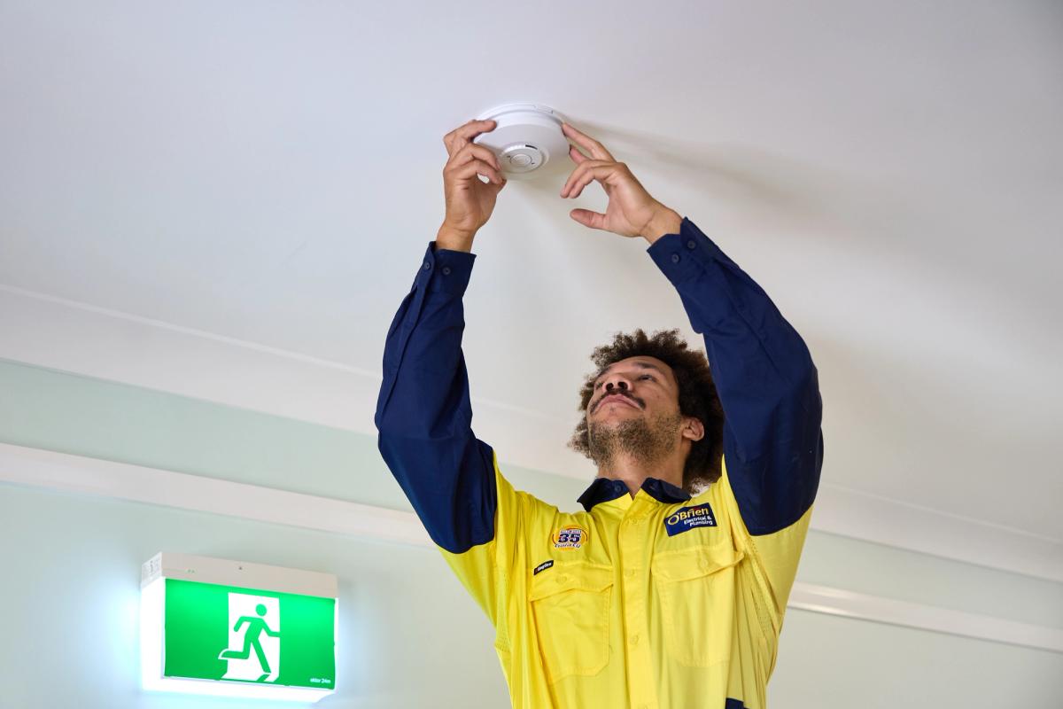 An O’Brien electrician fitting a smoke alarm to a ceiling beside an exit sign