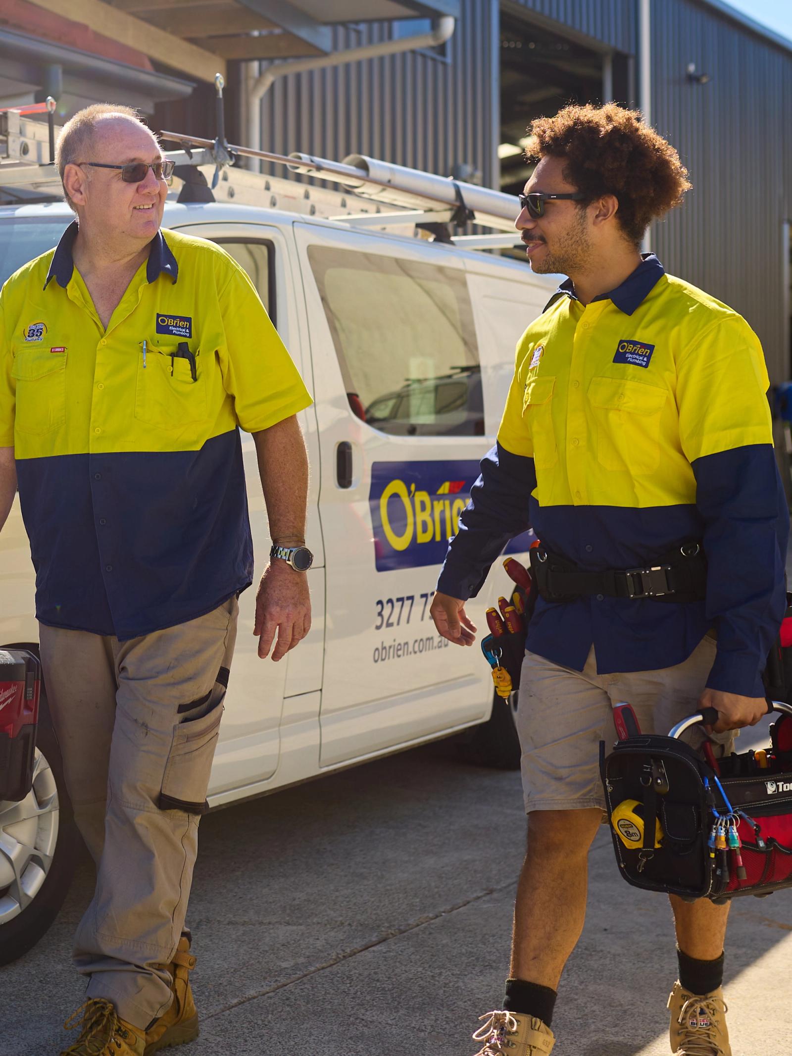 Two O’Brien technicians walking past a branch van with tool bags