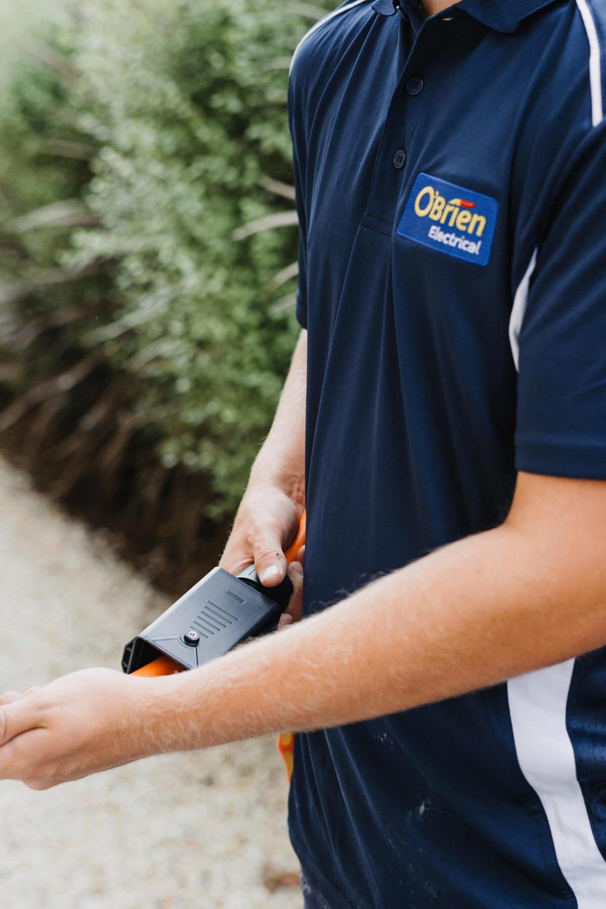 A technician in an O’Brien Electrical polo holding a tool beside a garden bed