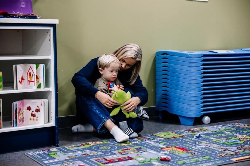 A woman embraces a toddler holding a green frog toy while sitting on a playmat in a classroom.