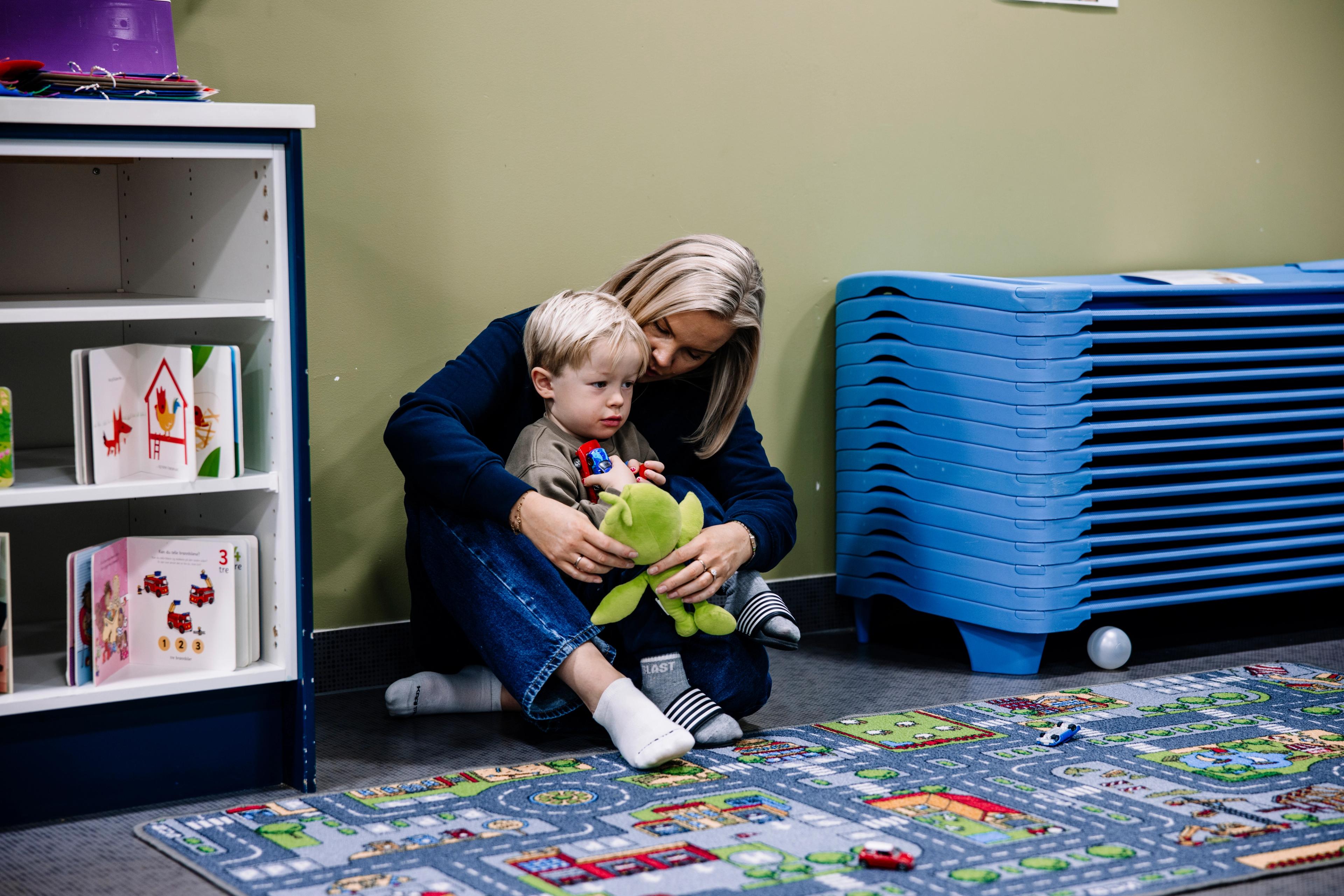 A woman embraces a toddler holding a green frog toy while sitting on a playmat in a classroom.