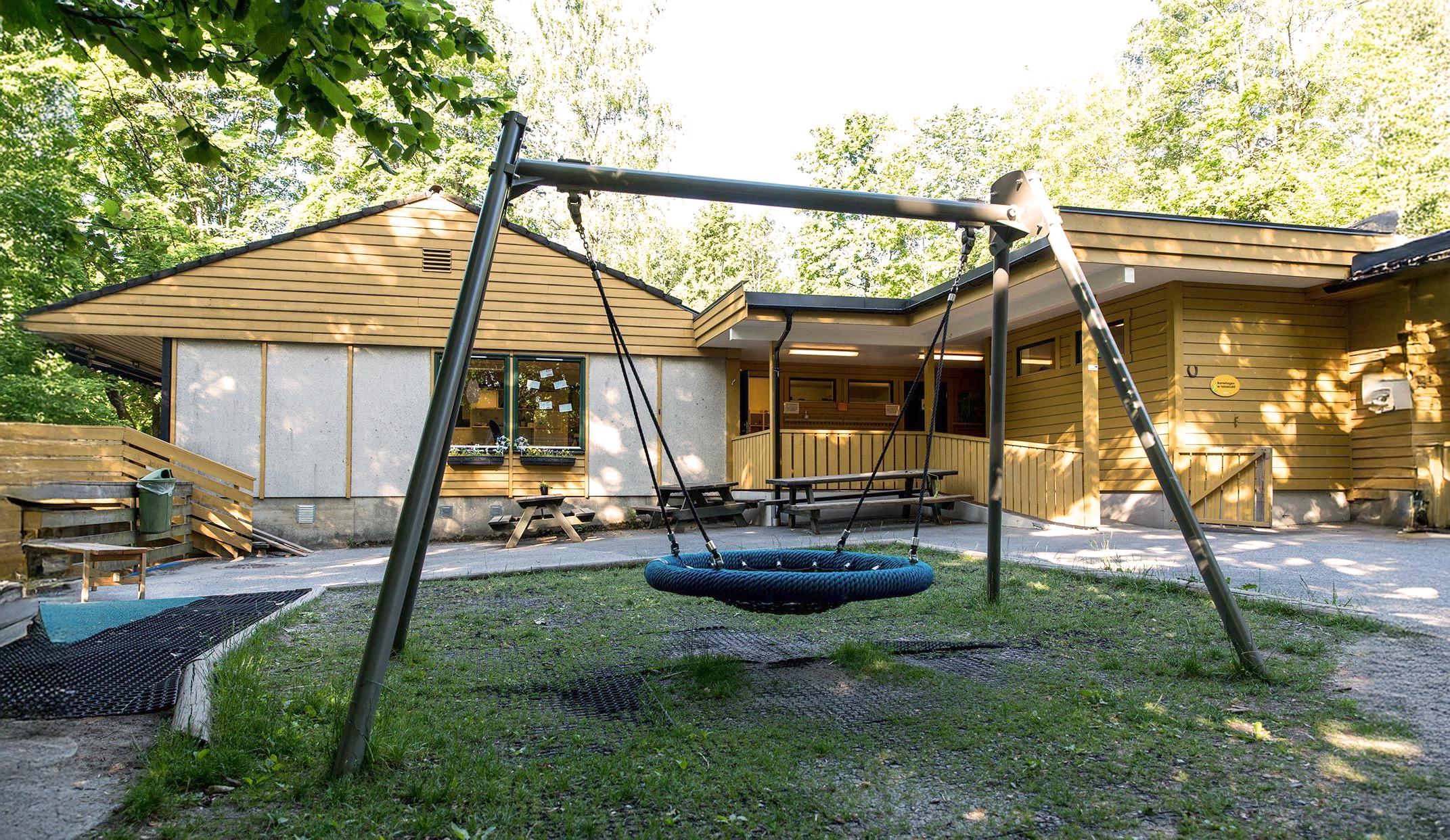 A playground with a large basket swing in front of a yellow wooden building surrounded by trees.