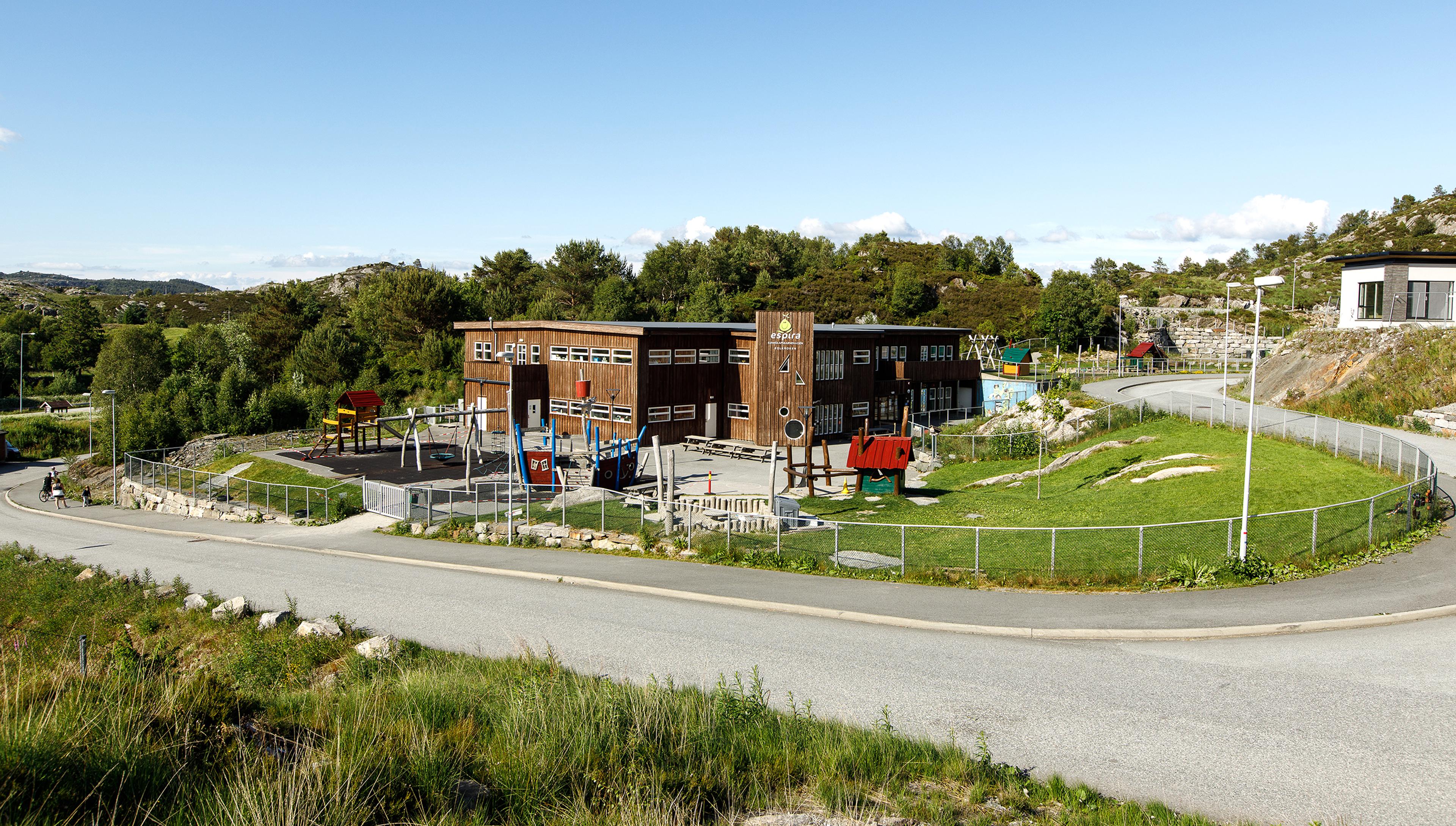 Fasaden på Espira Solkroken barnehage sett fra veien, med stort uteområde, grønne omgivelser og lekeapparater for barn