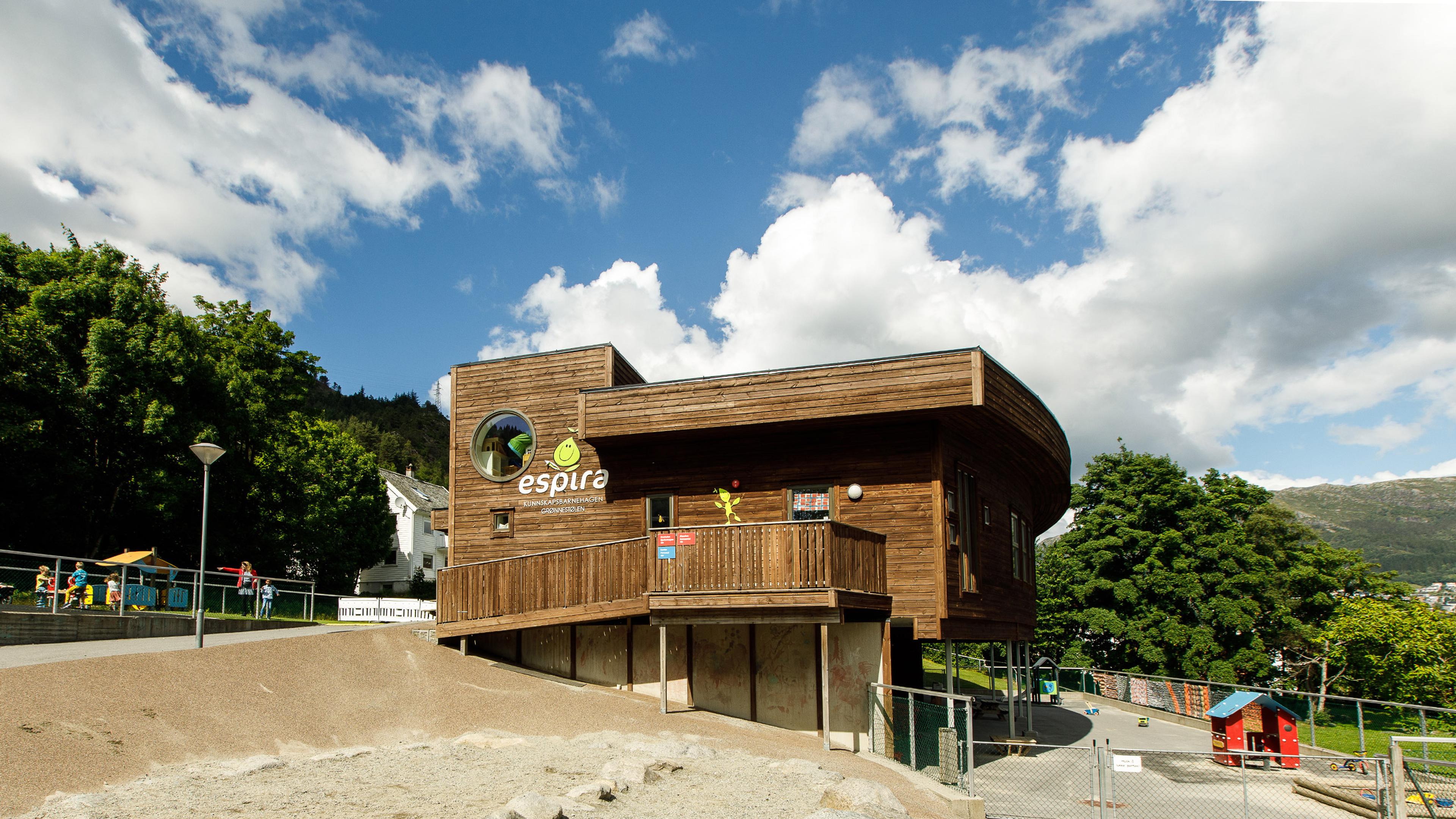 A modern, multi-level wooden building with a rounded facade and an "espira" logo, next to a playground under a blue sky with clouds.