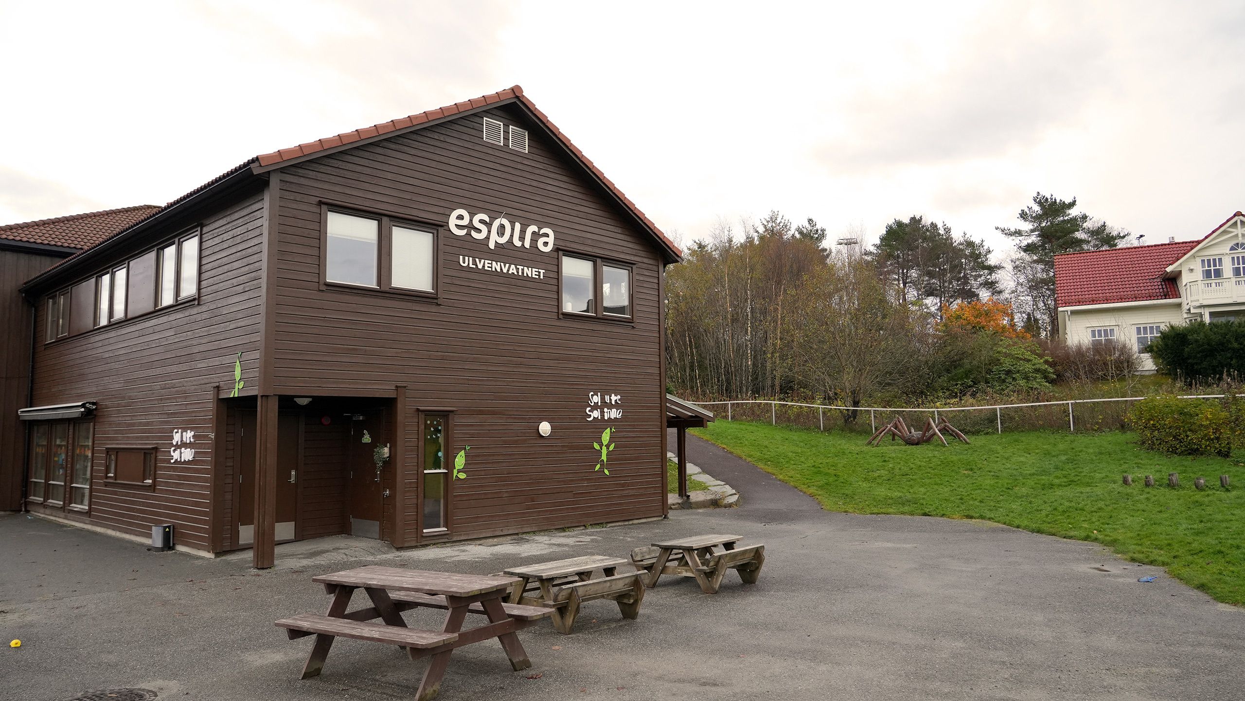 A brown wooden building with "espira ULVENVATNET" signage, picnic tables in the foreground, and a grassy area with trees and a house in the background.