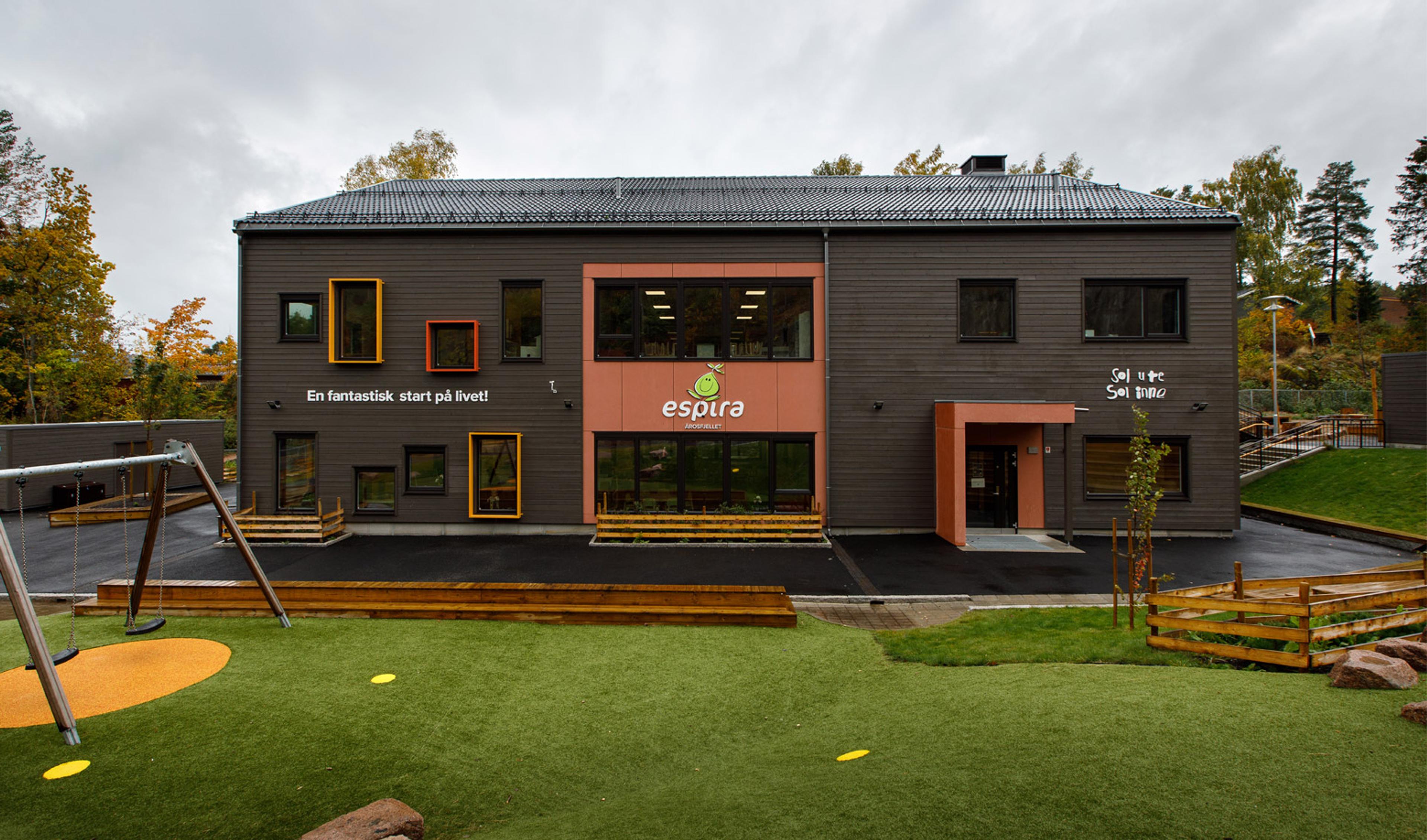 A modern dark-sided kindergarten building with colorful window frames, the Espira logo, and a playground with swings and artificial turf.