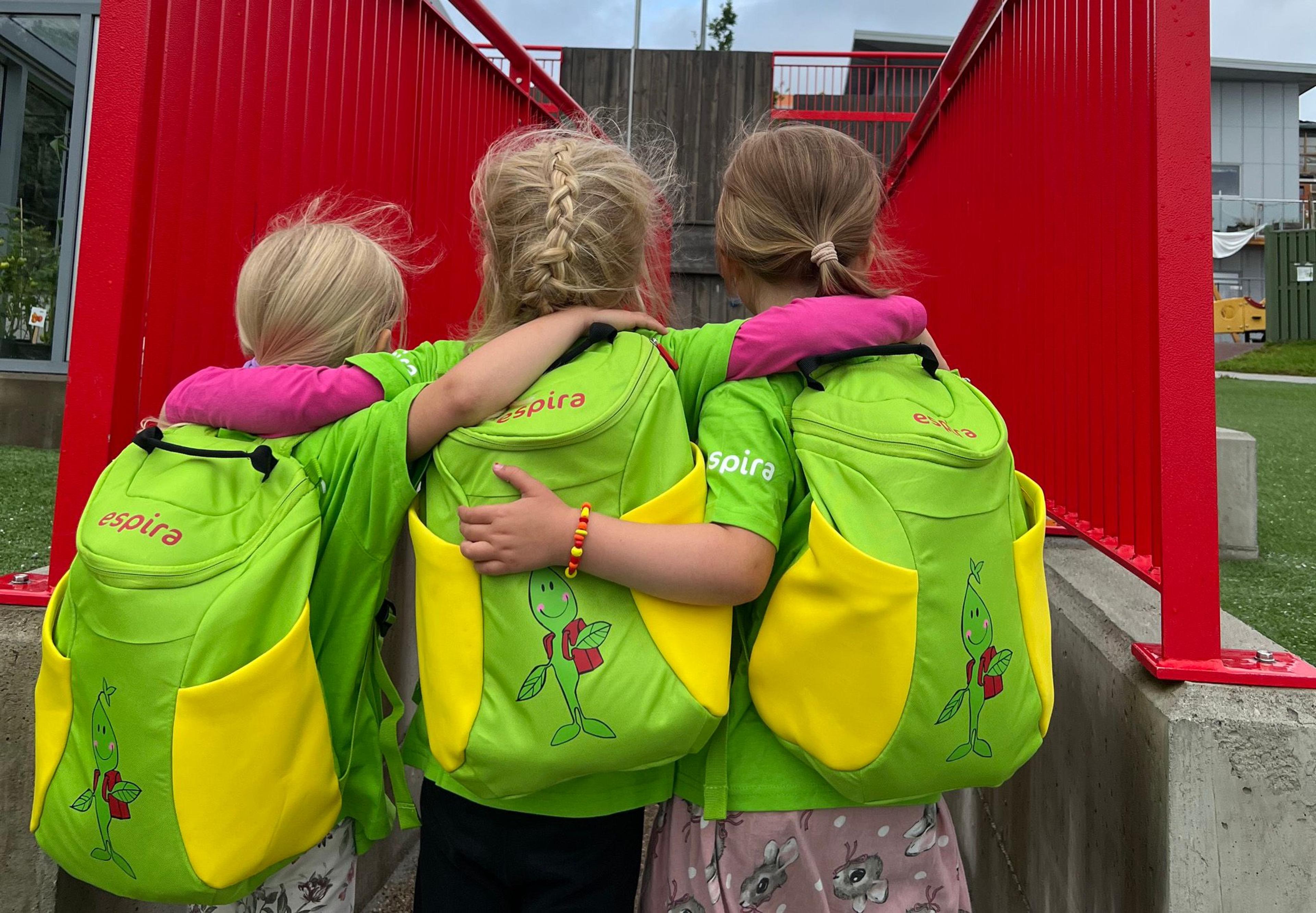 Tre barn med Skolespire-ryggsekker holder rundt hverandre, med ryggen til kamera.