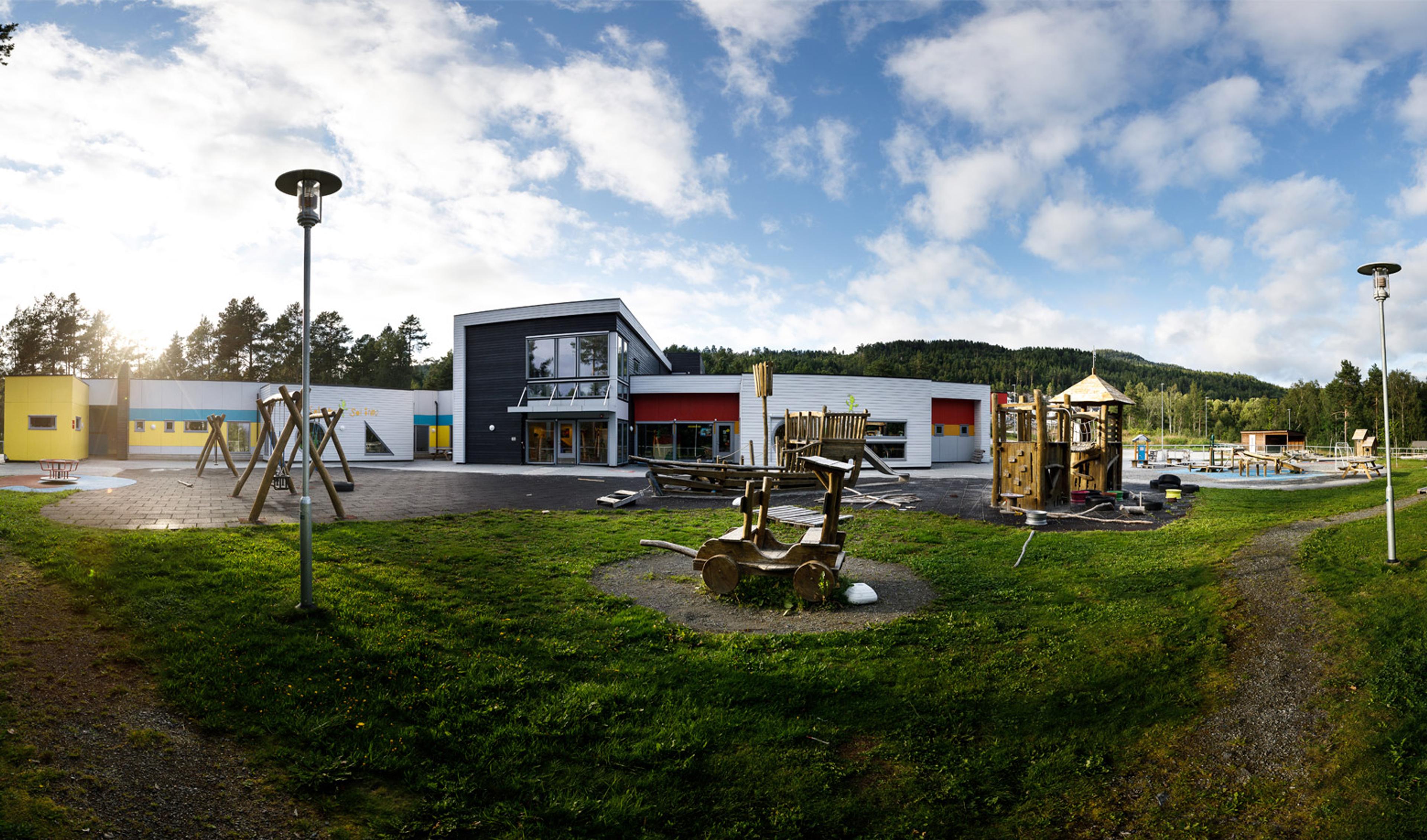 Modern kindergarten building with a colorful facade and a large outdoor playground featuring swings and wooden structures.