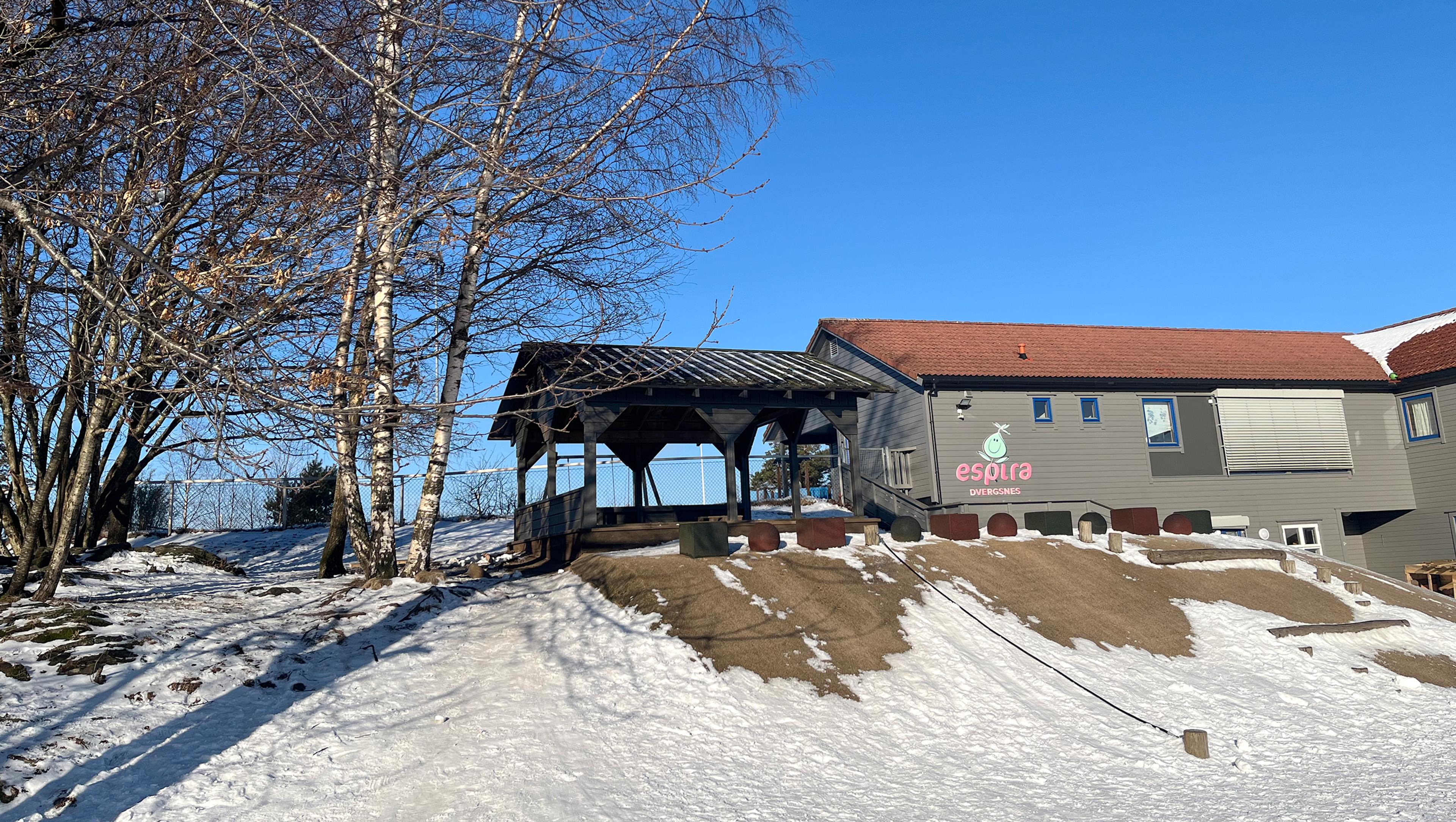 A grey building with a red roof and the 'Espira Barnehager' logo, a covered walkway, bare trees, and patchy snow on the ground.