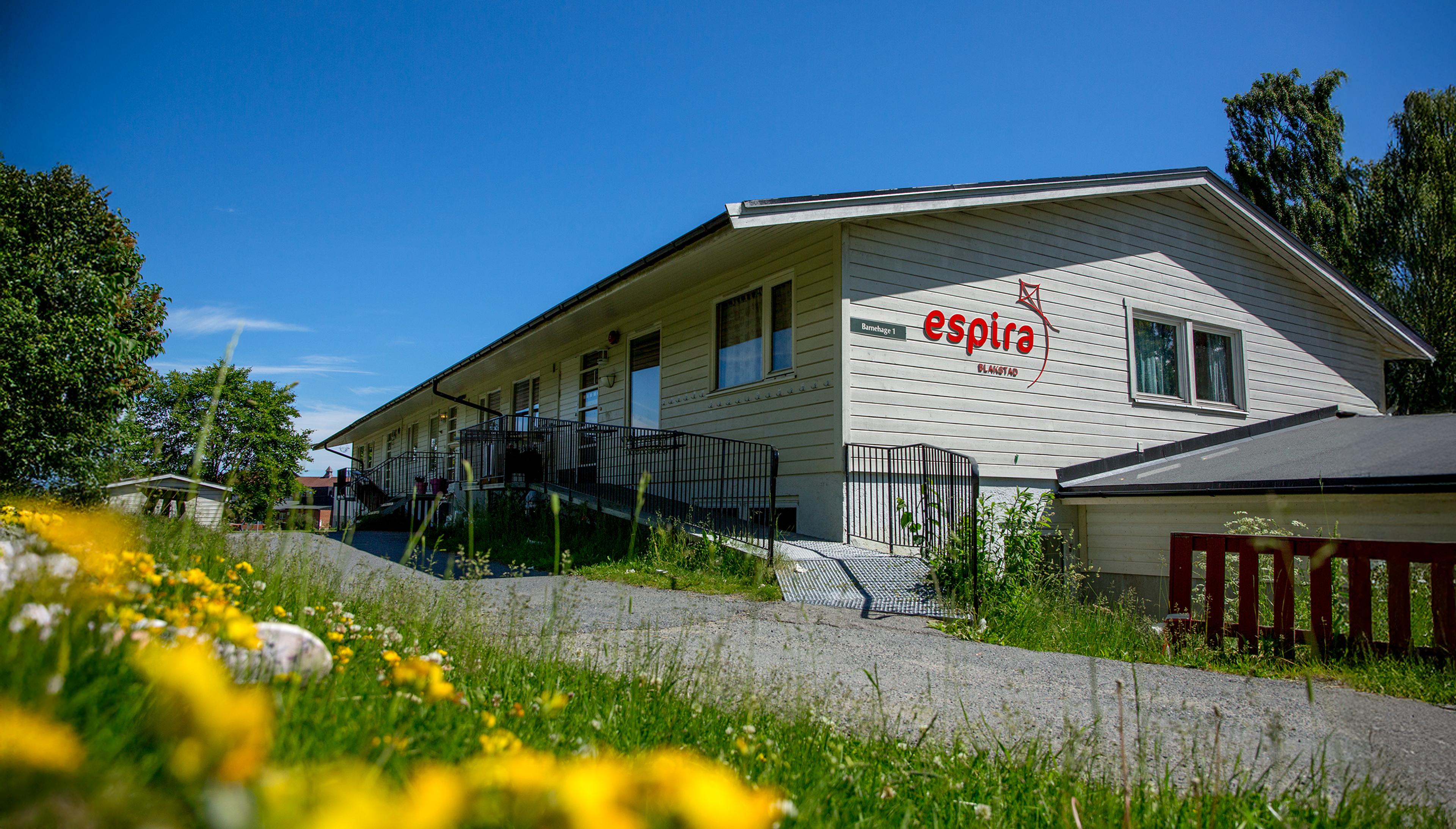 A light-colored building with an "espira" logo and an accessible ramp, surrounded by green grass and yellow flowers under a clear blue sky.