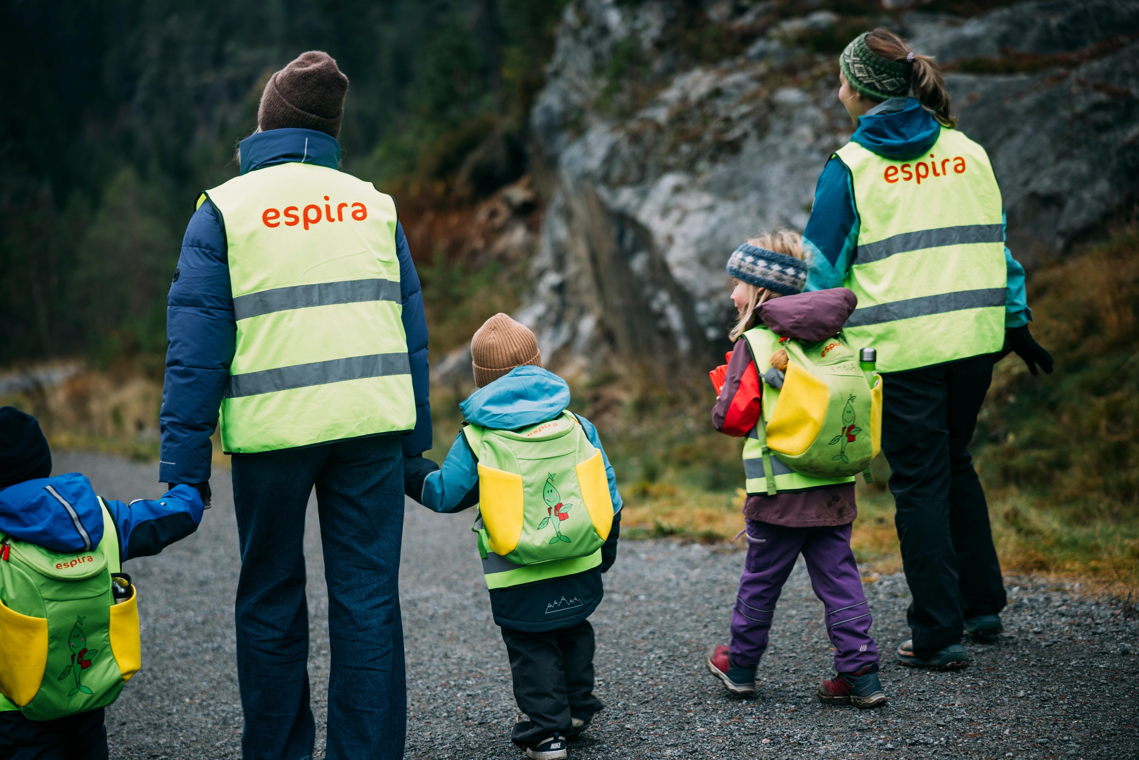 To voksne i refleksvester med «Espira»-logo og tre barn med refleksvester og ryggsekker går bortover en utendørs sti.