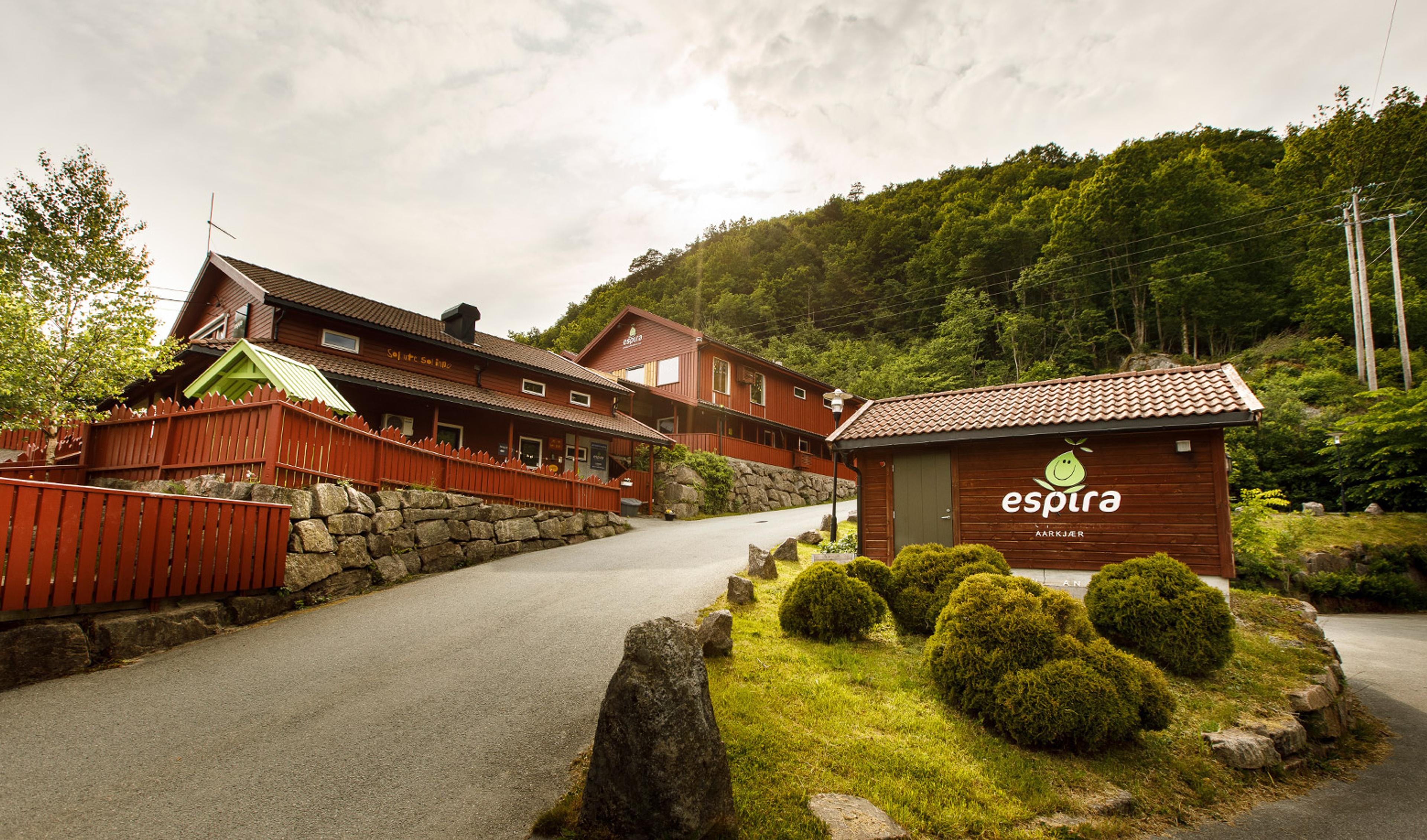 A paved road winds past red wooden buildings and a smaller building with an "Espira" logo, set against a green forested hillside.