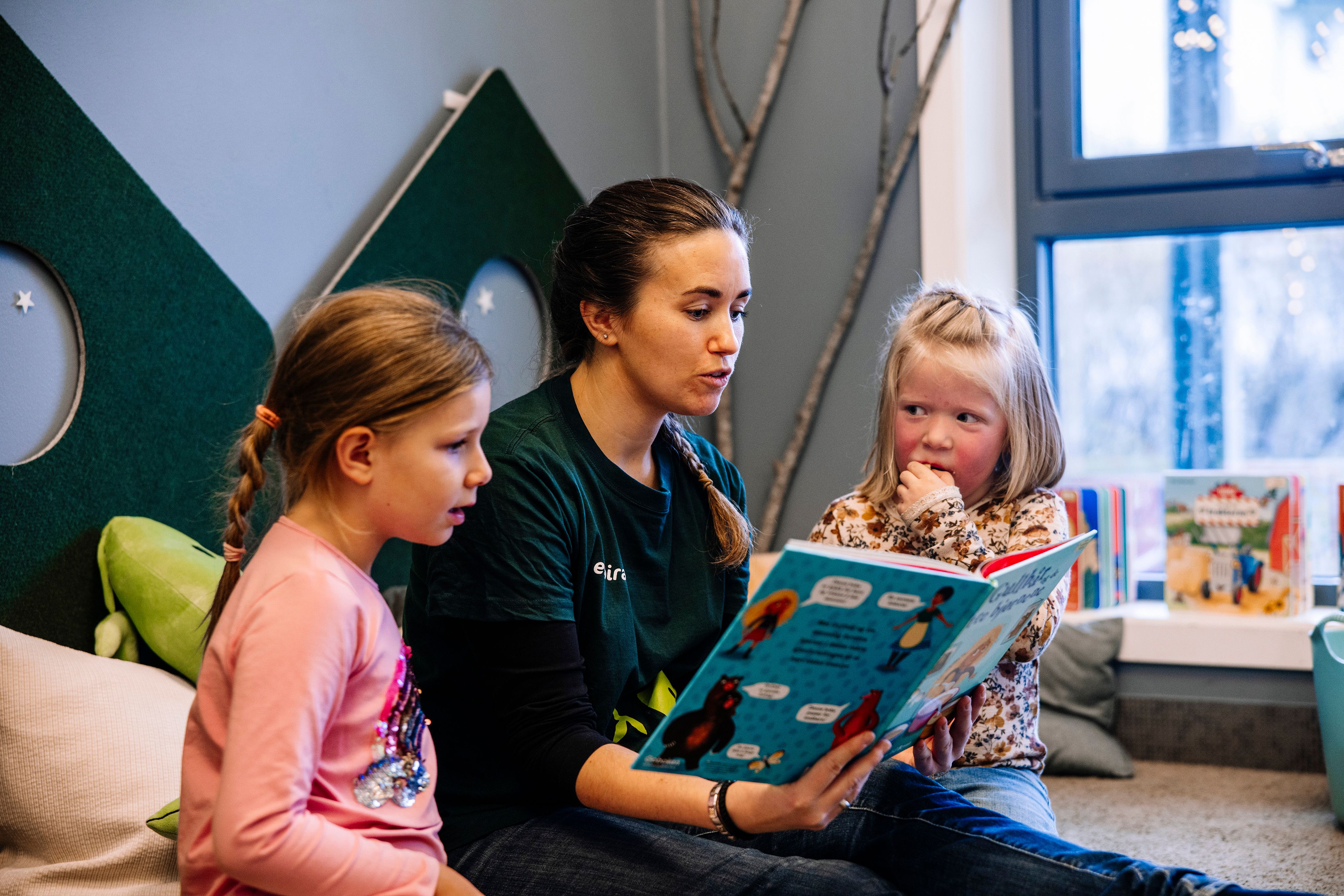 Barnehageansatt leser i bok for to barn, i et rom med puter og store vinduer.