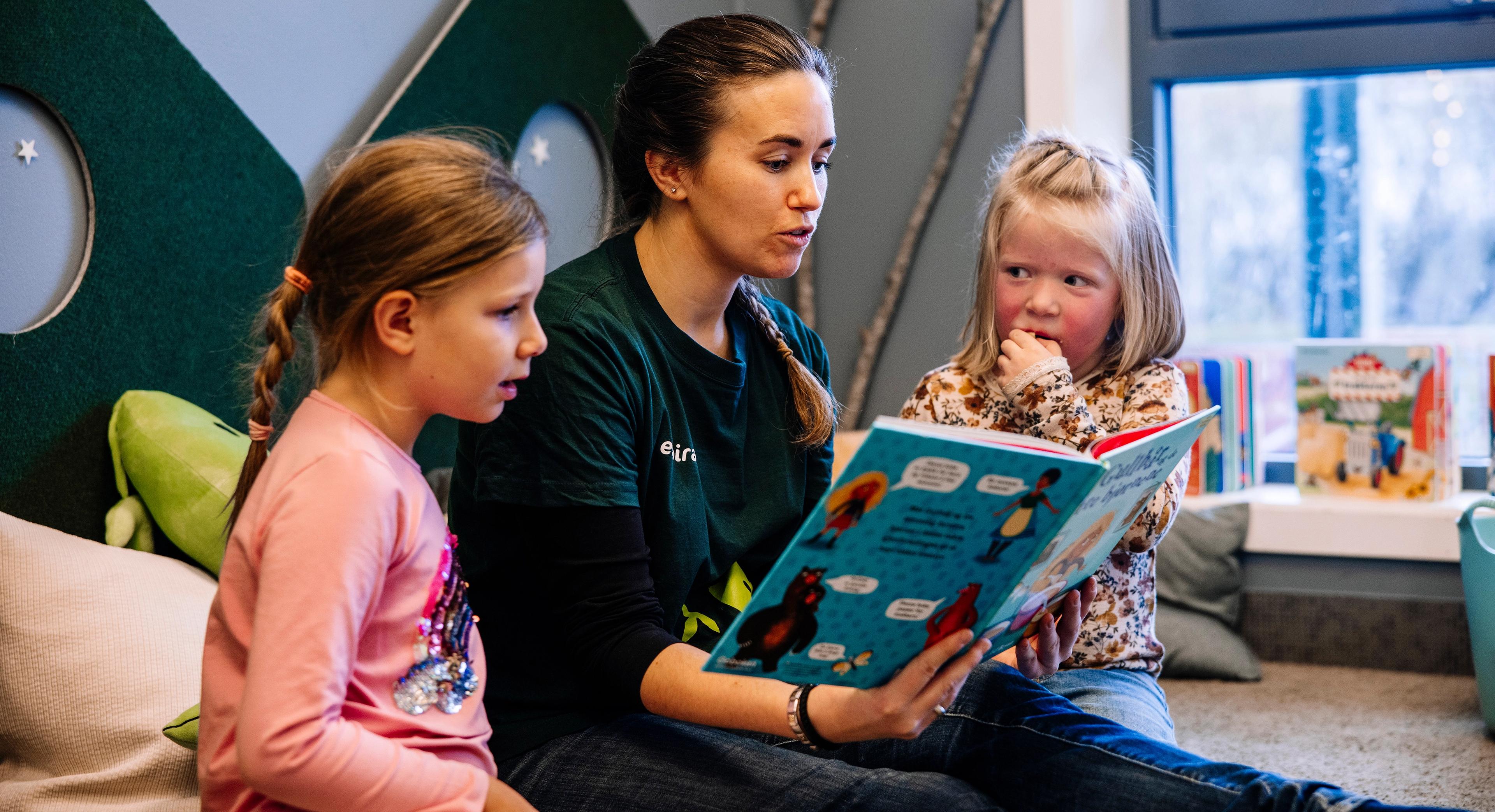 Barnehageansatt leser i bok for to barn, i et rom med puter og store vinduer.