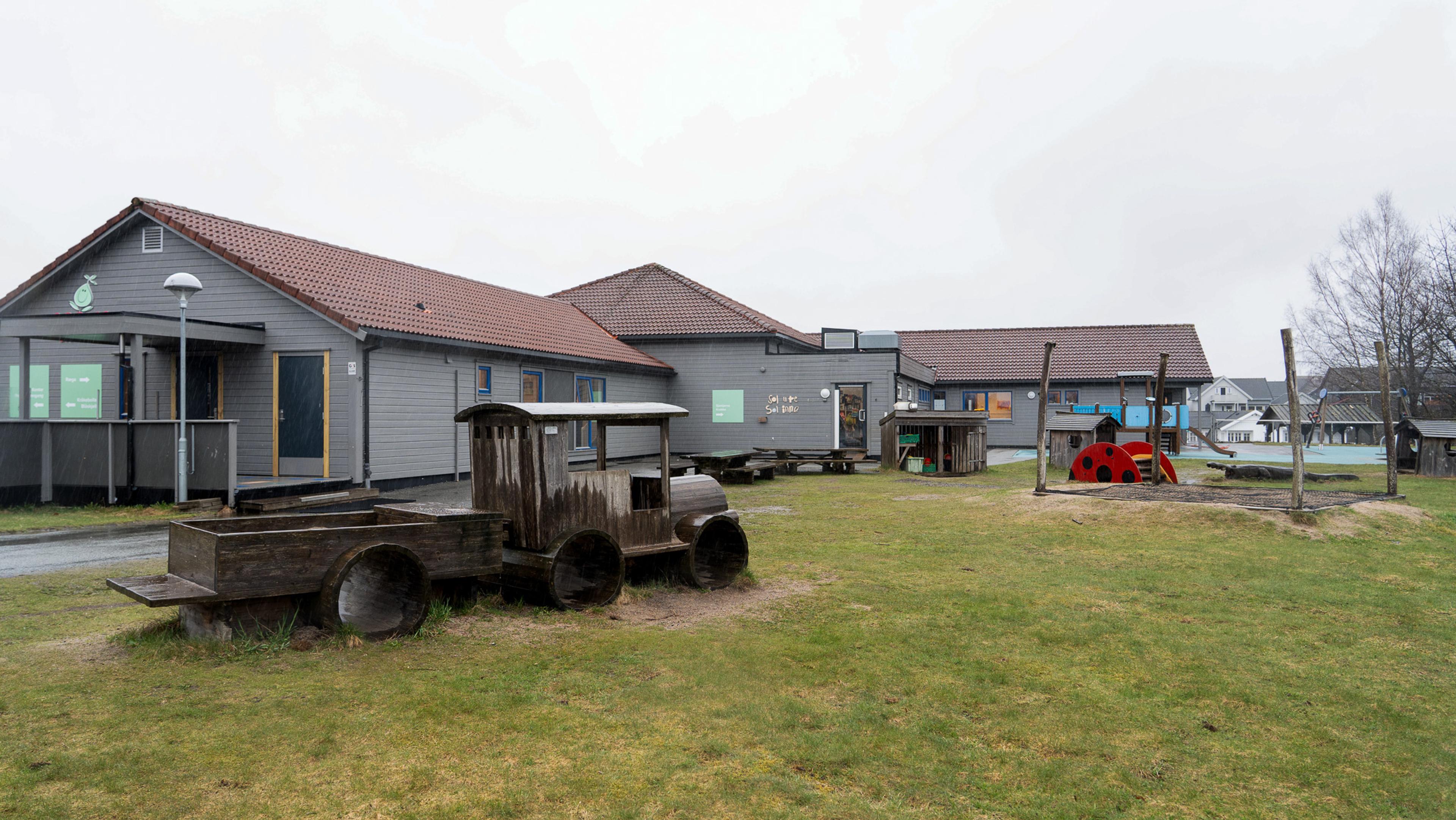 A grey building with a red roof stands next to a grassy playground featuring a wooden train and various play structures.