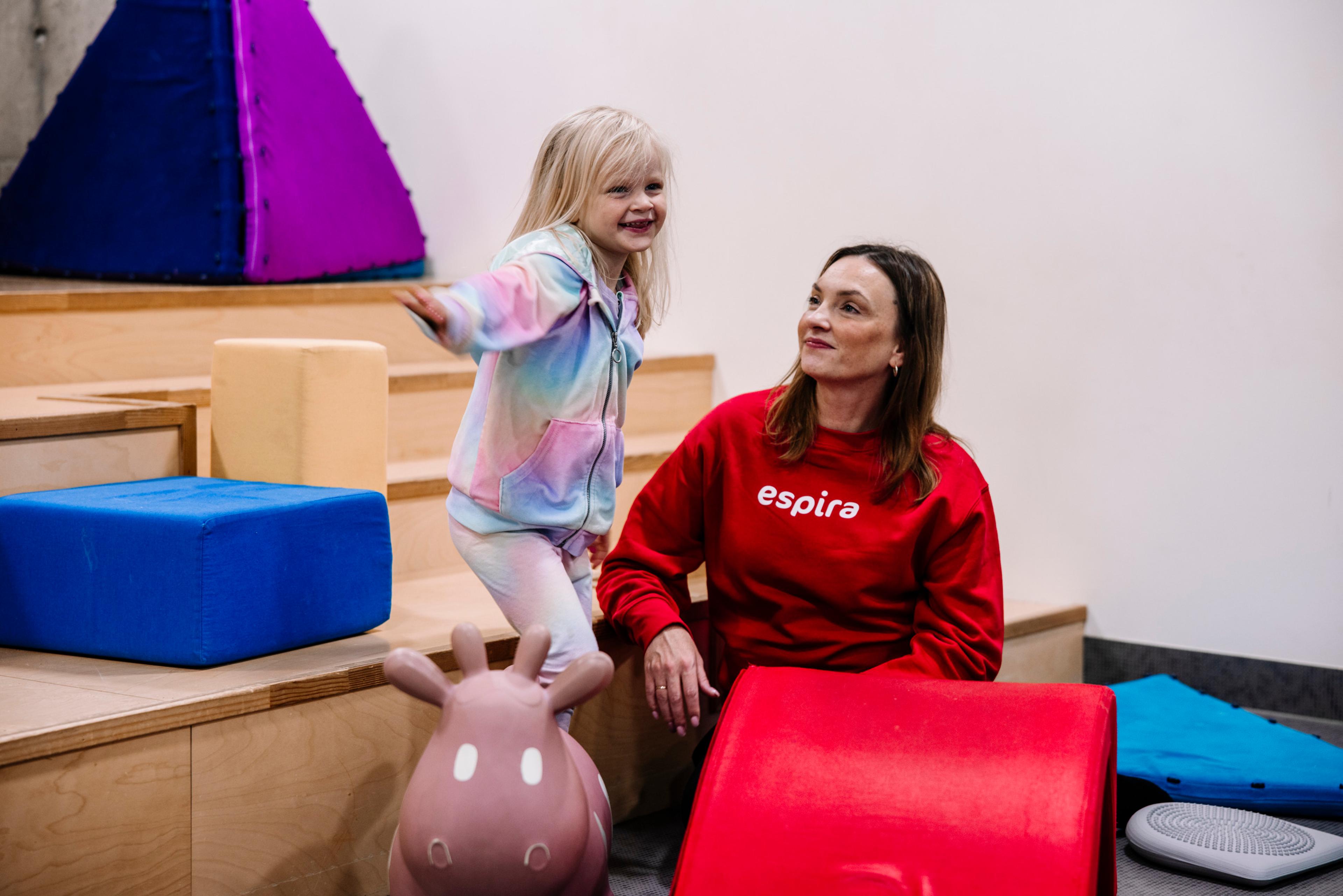 En barnehageansatt med Espira-genser smiler mot et barn som balanserer på en pute.