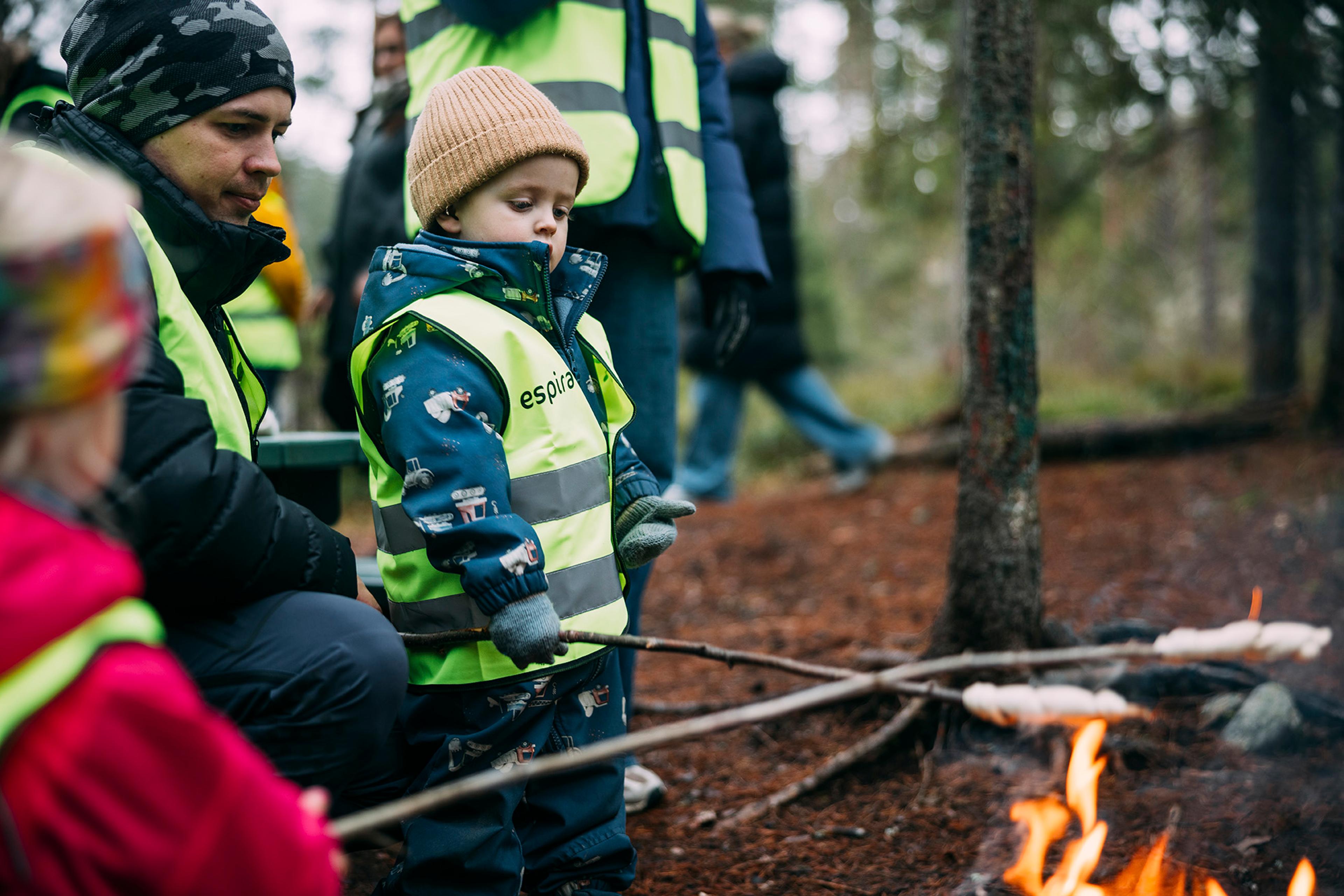 Barn griller pinnebrød rundt et bål i skogen.