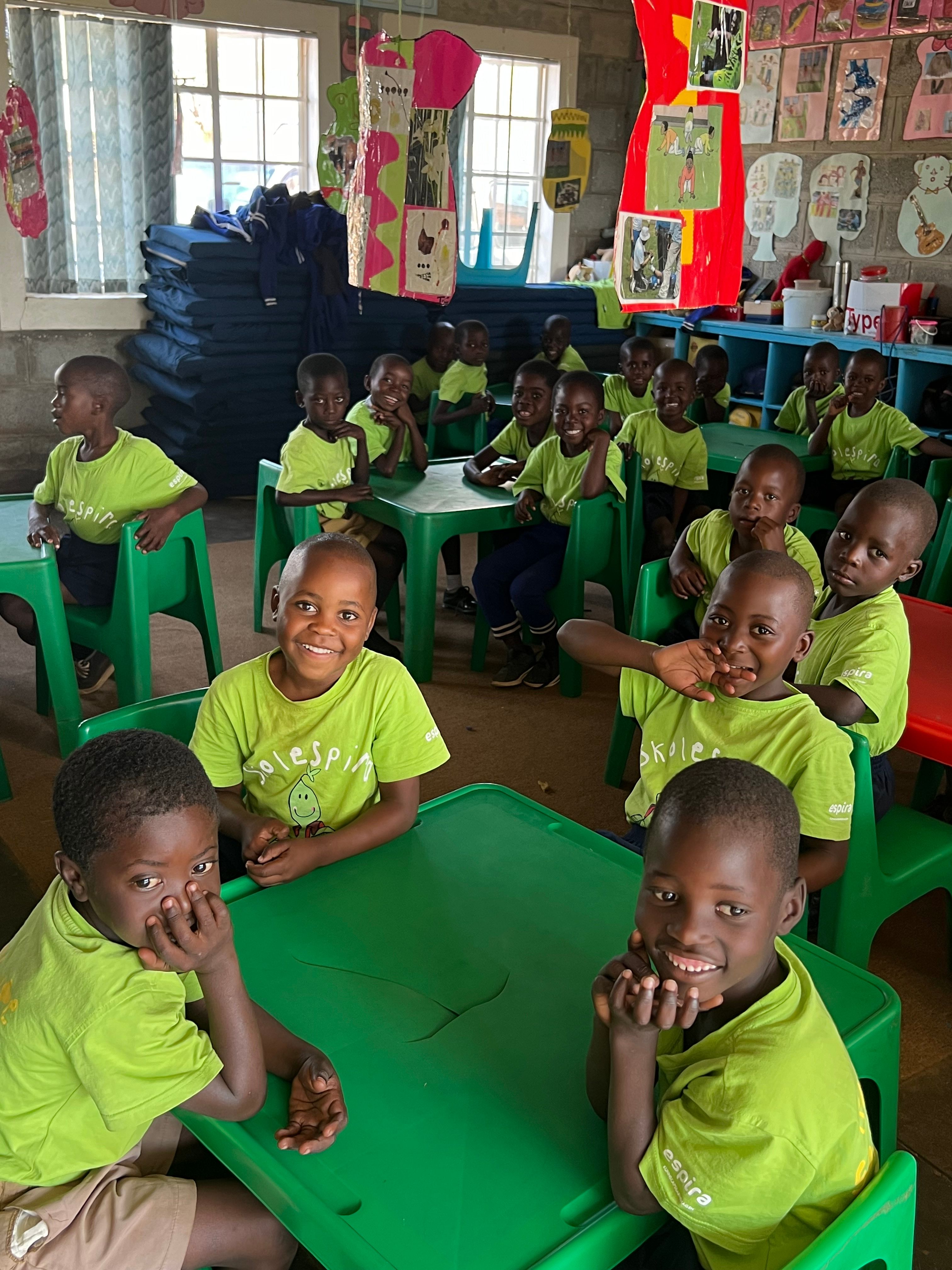 A group of smiling children in bright green t-shirts sit at green tables in a classroom.