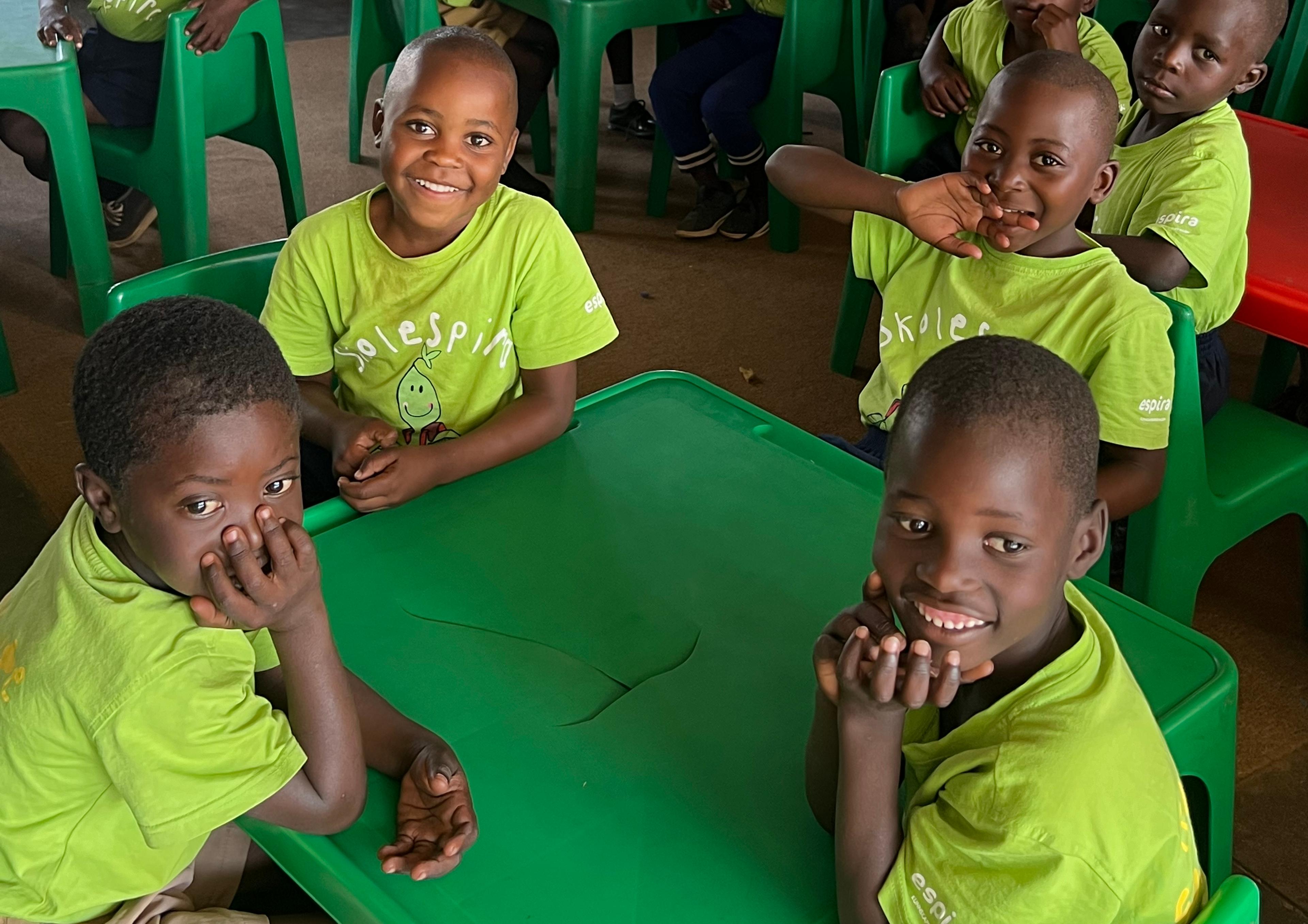 A group of smiling children in bright green t-shirts sit at green tables in a classroom.