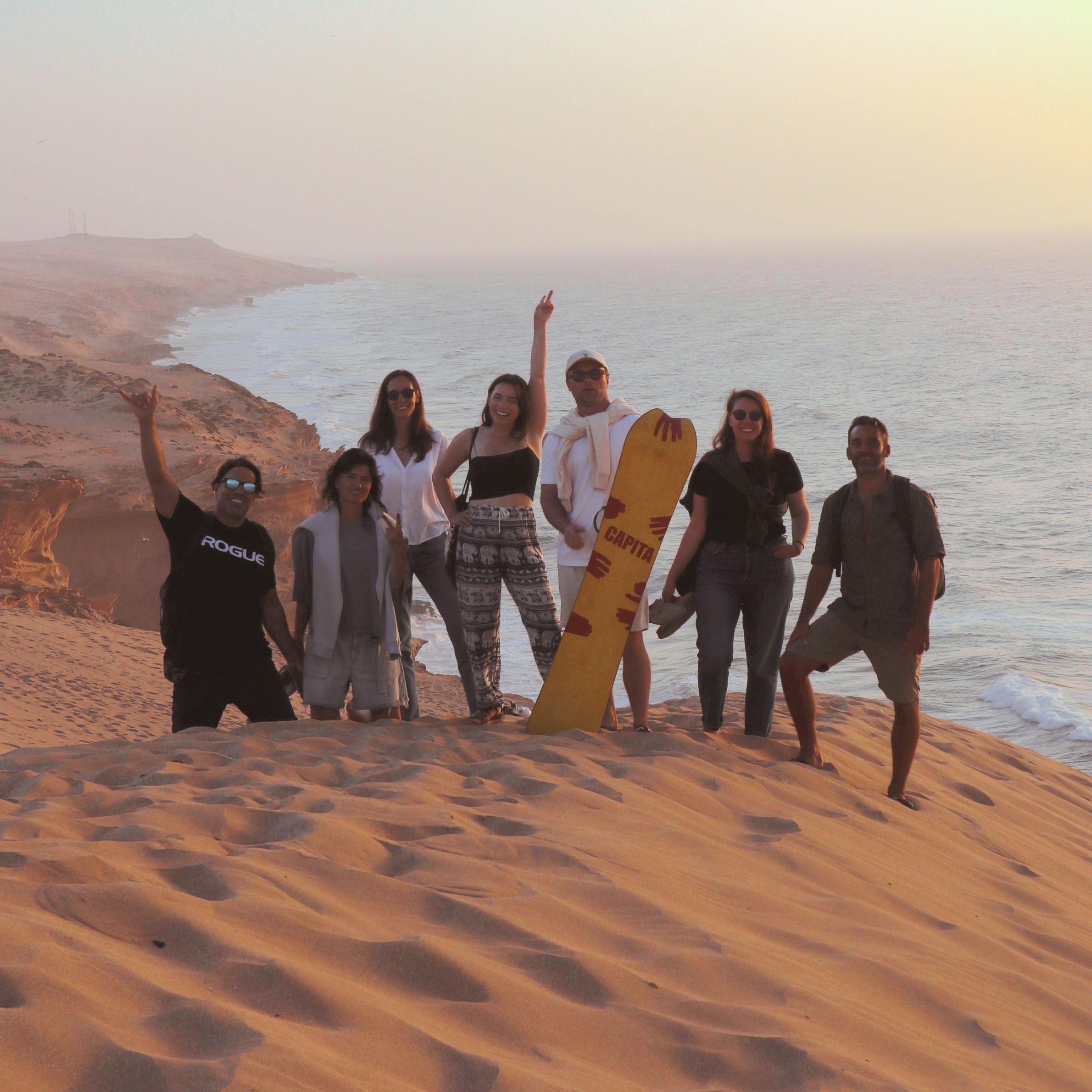 Group of friends celebrating on the desert dunes at sunset during a Sea Me Smile surf trip