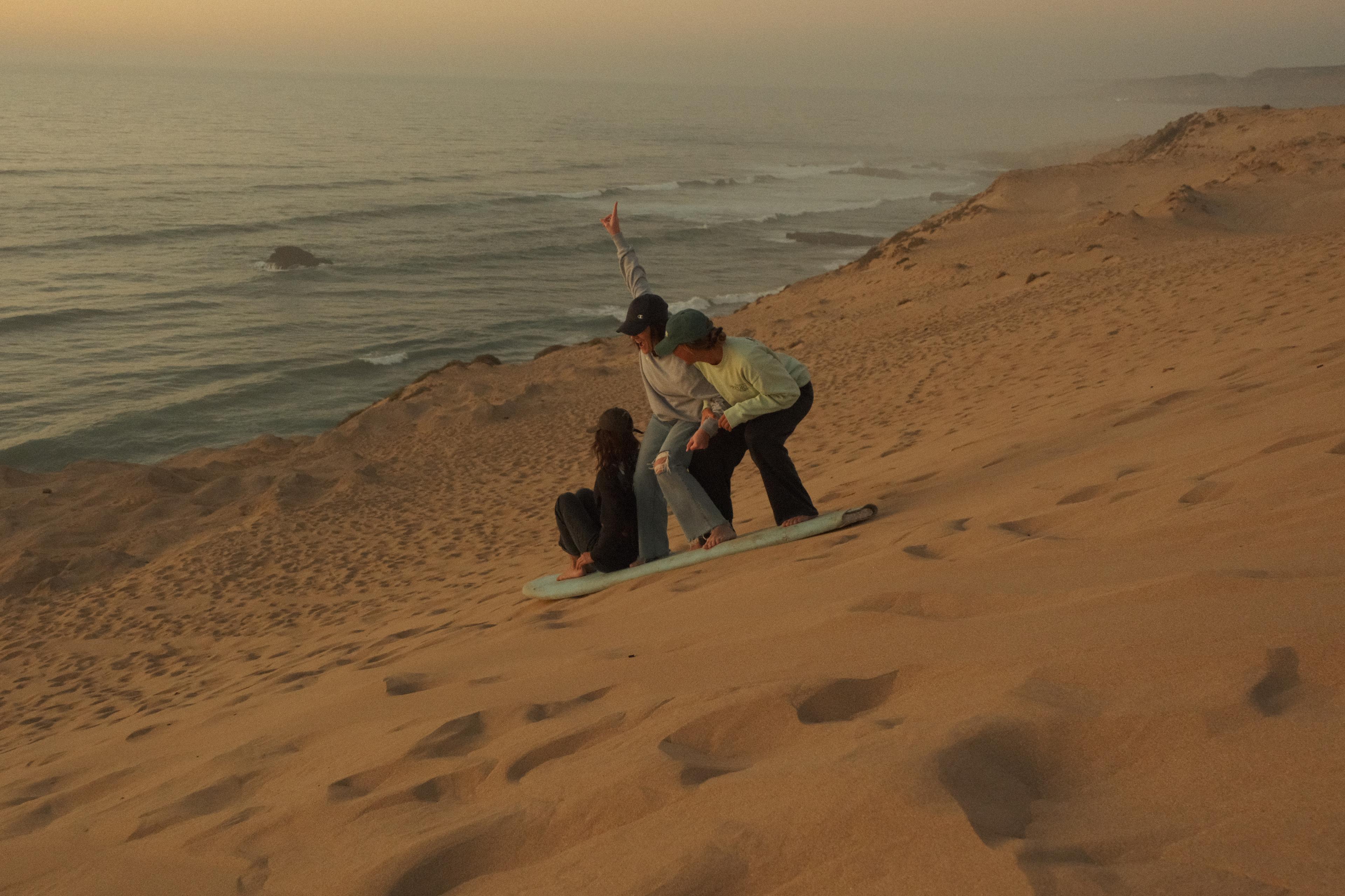Friends sandboarding down a desert dune at sunset, Morocco