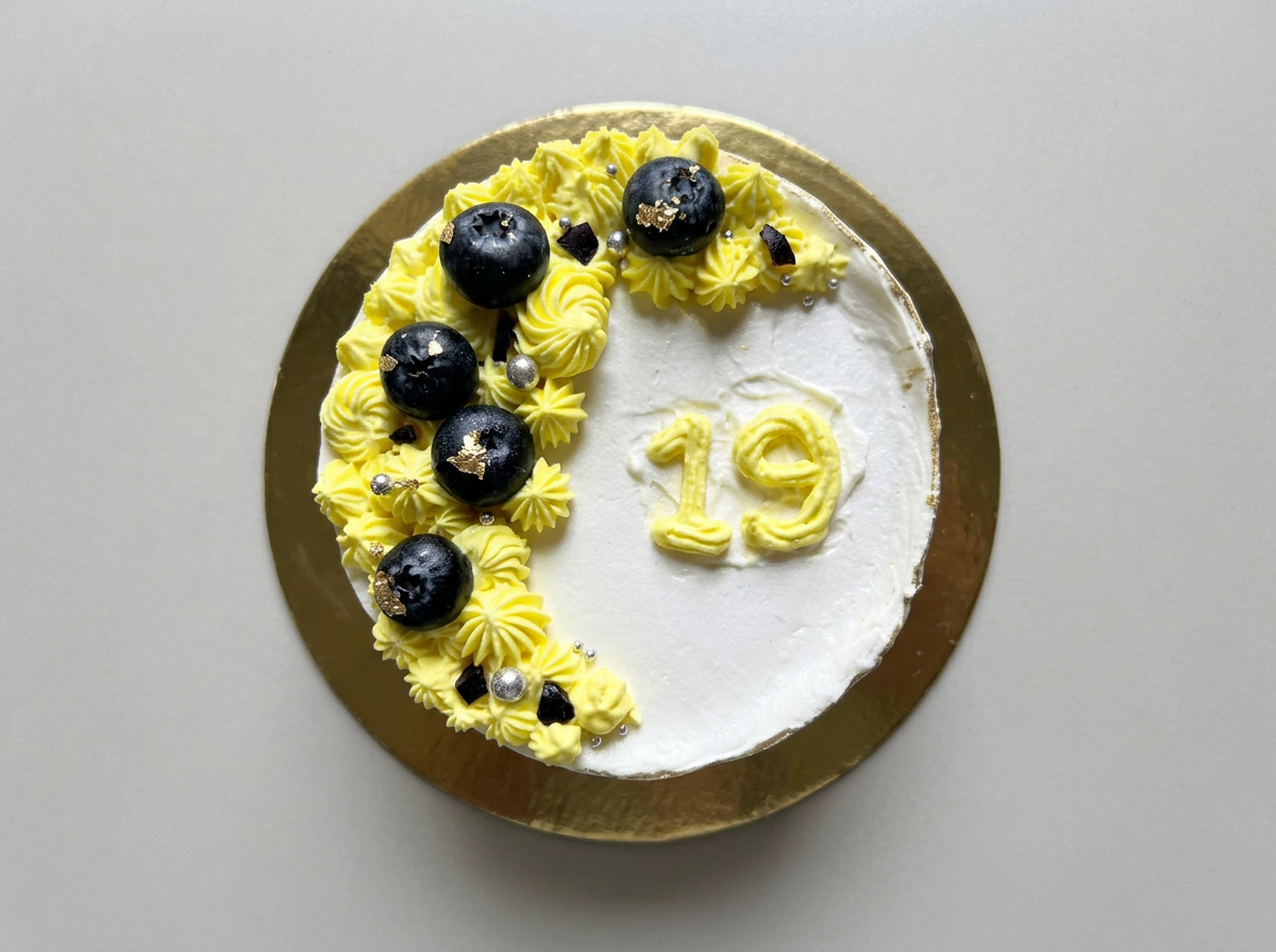 White cake with a crescent of lemon-yellow buttercream rosettes, fresh blueberries and a piped number nineteen