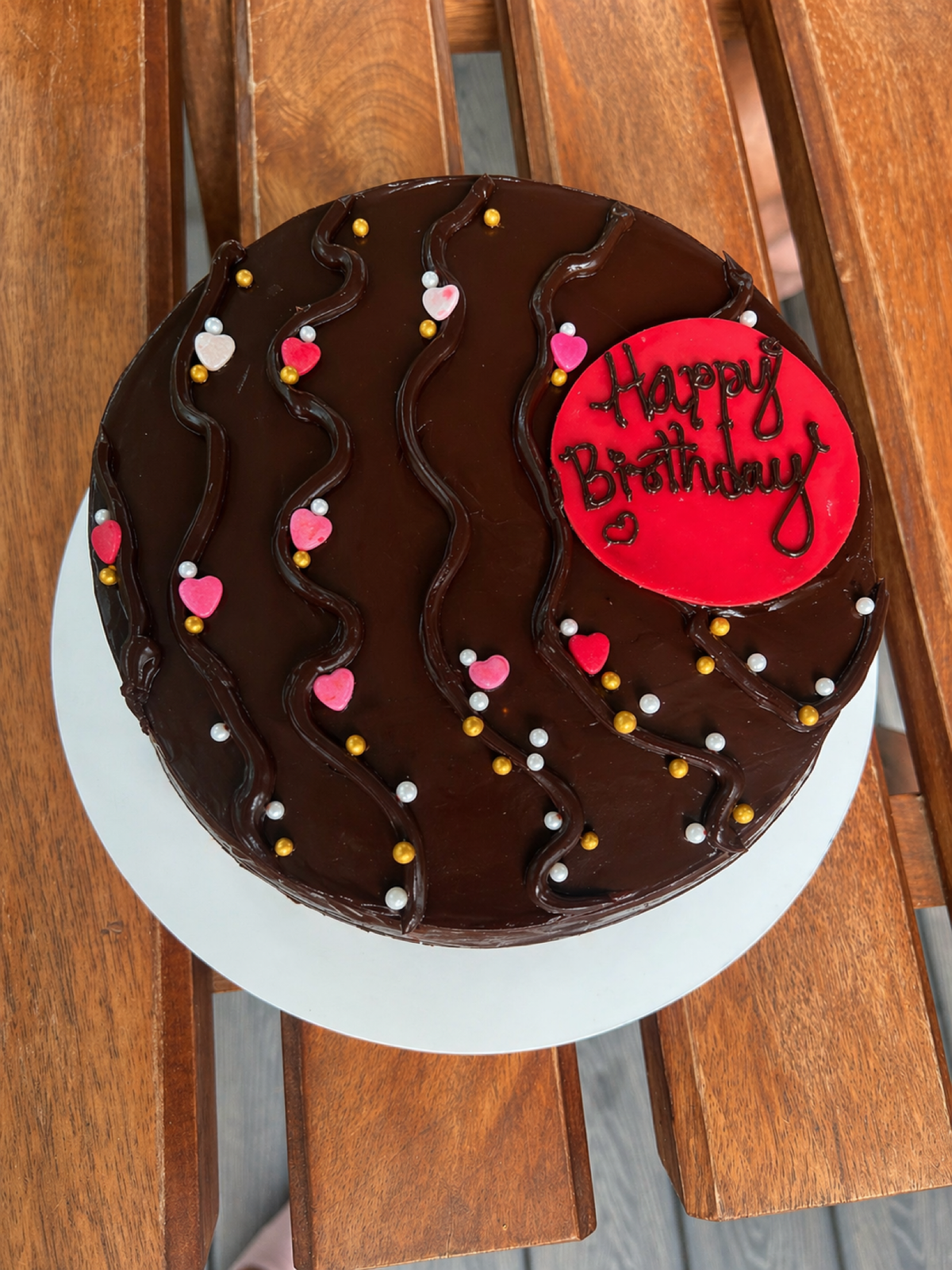 Top-down chocolate ganache cake decorated with pink candy hearts, gold pearls and a red Happy Birthday plaque