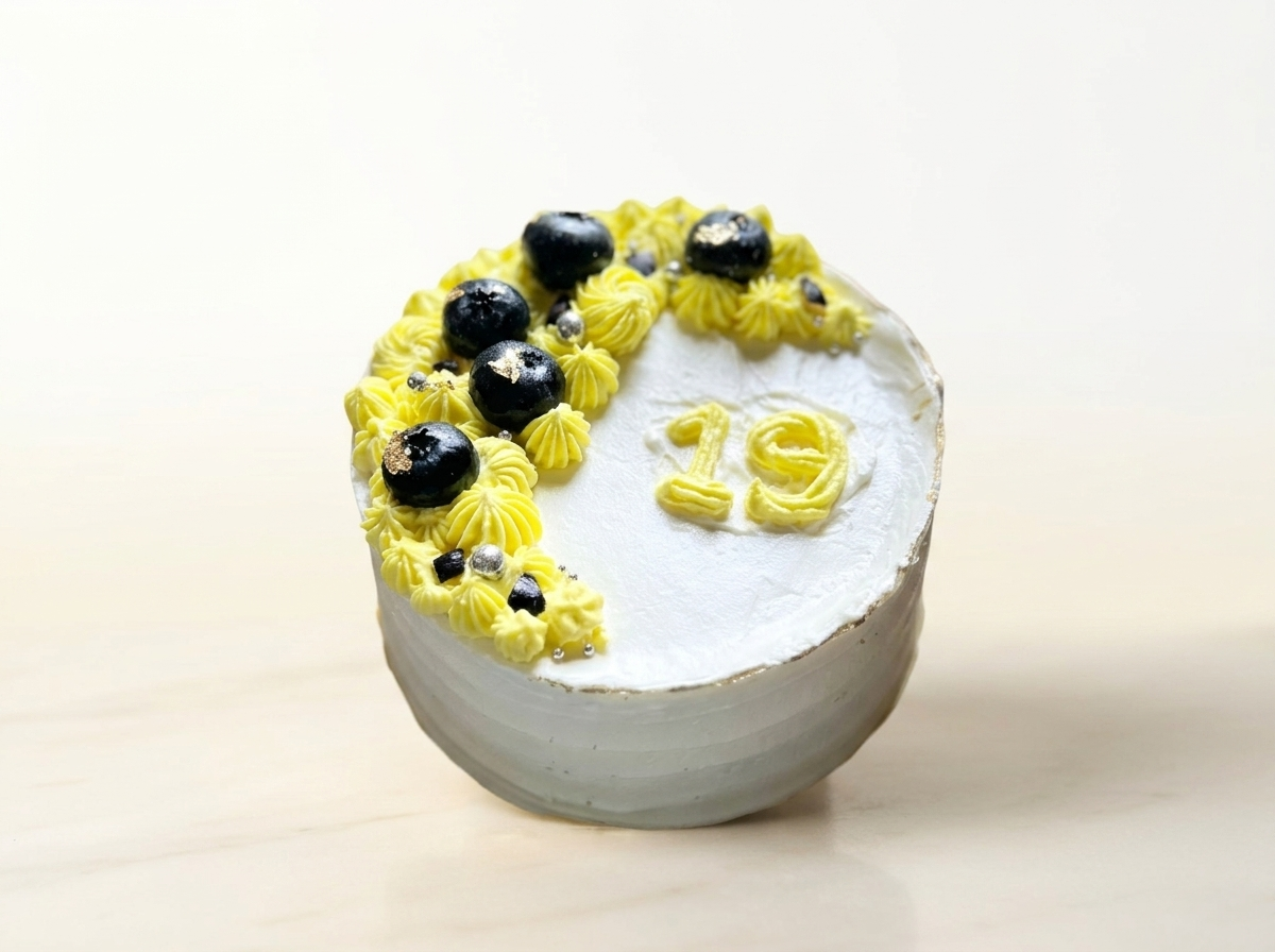 White cake with a crescent of lemon-yellow buttercream rosettes, fresh blueberries and a piped number nineteen