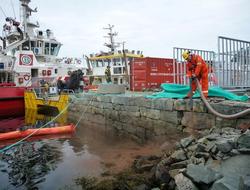 Mann i arbeidsklær holder slange ved bryggekanten nær to båter og en container. Overskyet vær. Fotografi.