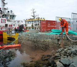 Mann i arbeidsklær holder slange ved bryggekanten nær to båter og en container. Overskyet vær. Fotografi.