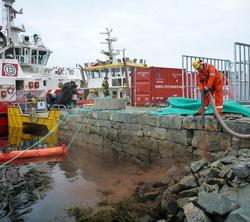 Mann i arbeidsklær holder slange ved bryggekanten nær to båter og en container. Overskyet vær. Fotografi.